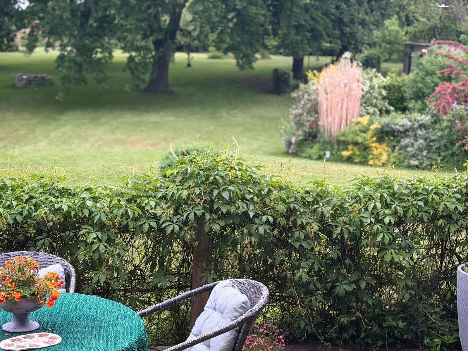 Gartenansicht mit grünem Rasen, Bäumen im Hintergrund, bunten Blumen und Gartenmöbeln im Vordergrund.