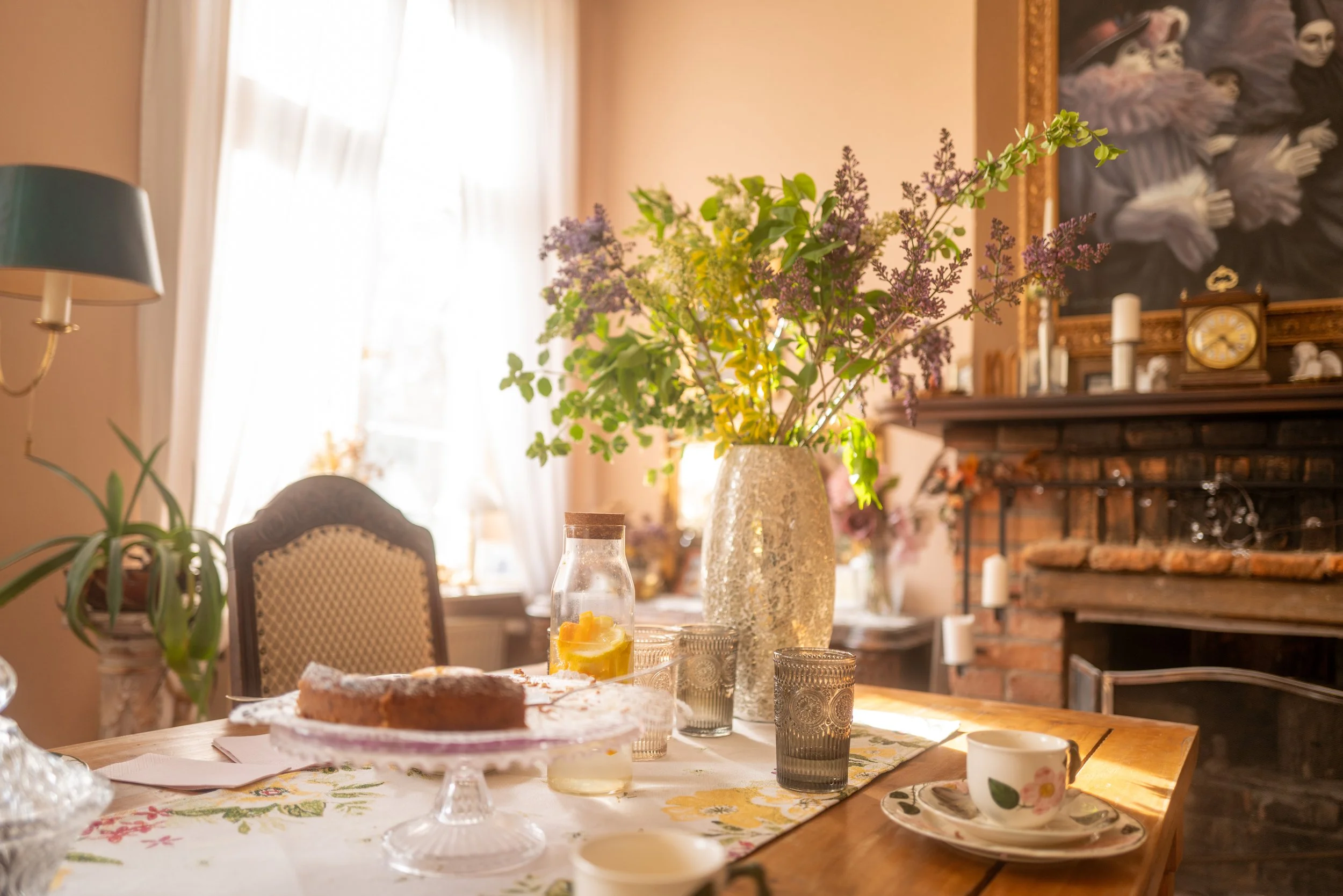 Ein festlich gedeckter Esstisch mit einem Blumenstrauß in einer Vase, Kuchen, Getränken und Geschirr in einem gemütlichen Wohnzimmer mit Kamin und Kunstwerken an der Wand.