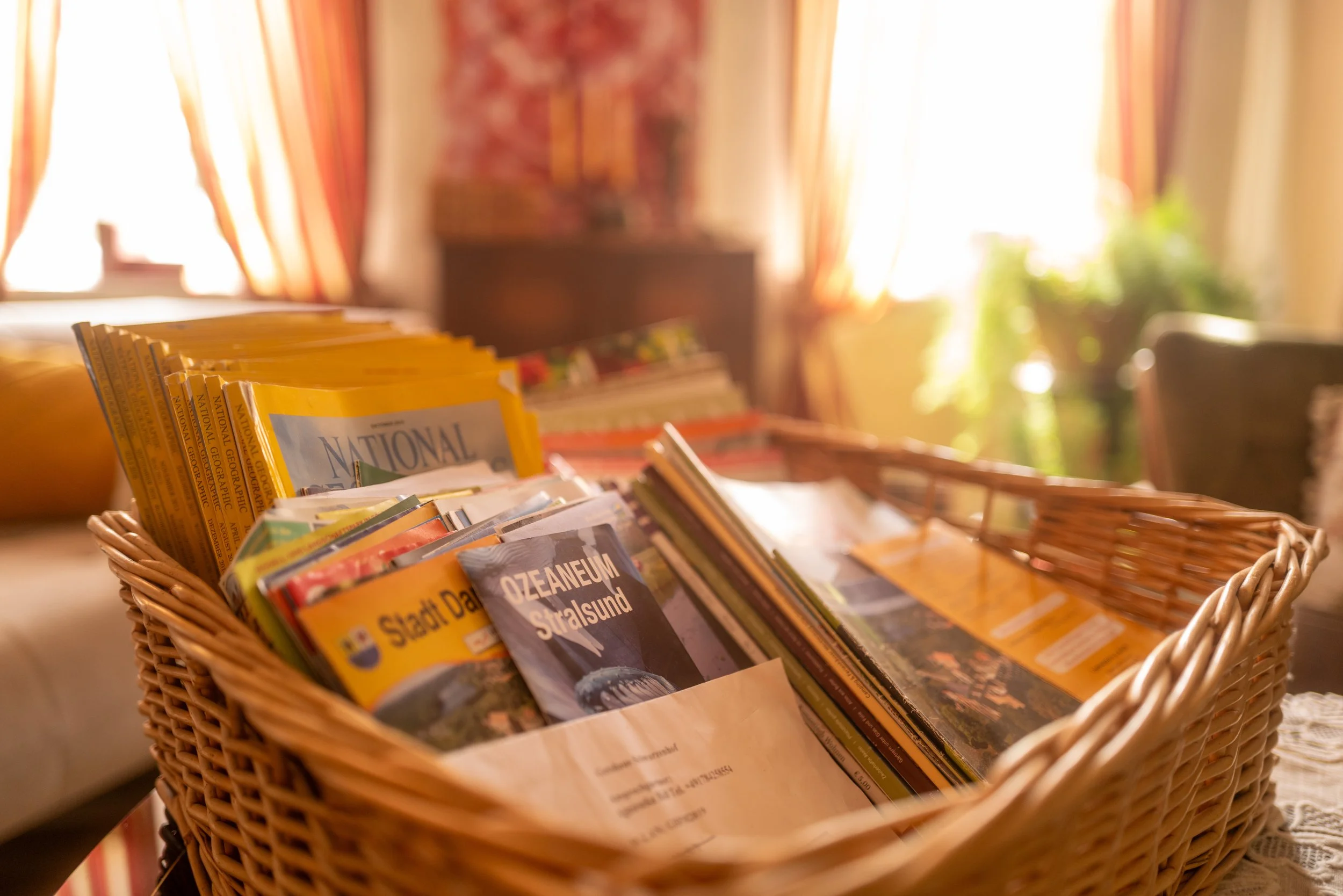 Ein Korb voller Zeitschriften und Magazine auf einem Tisch, mit einem Hintergrund aus Vorhängen und Pflanzen, die Raum mit natürlichem Licht durchfluten.