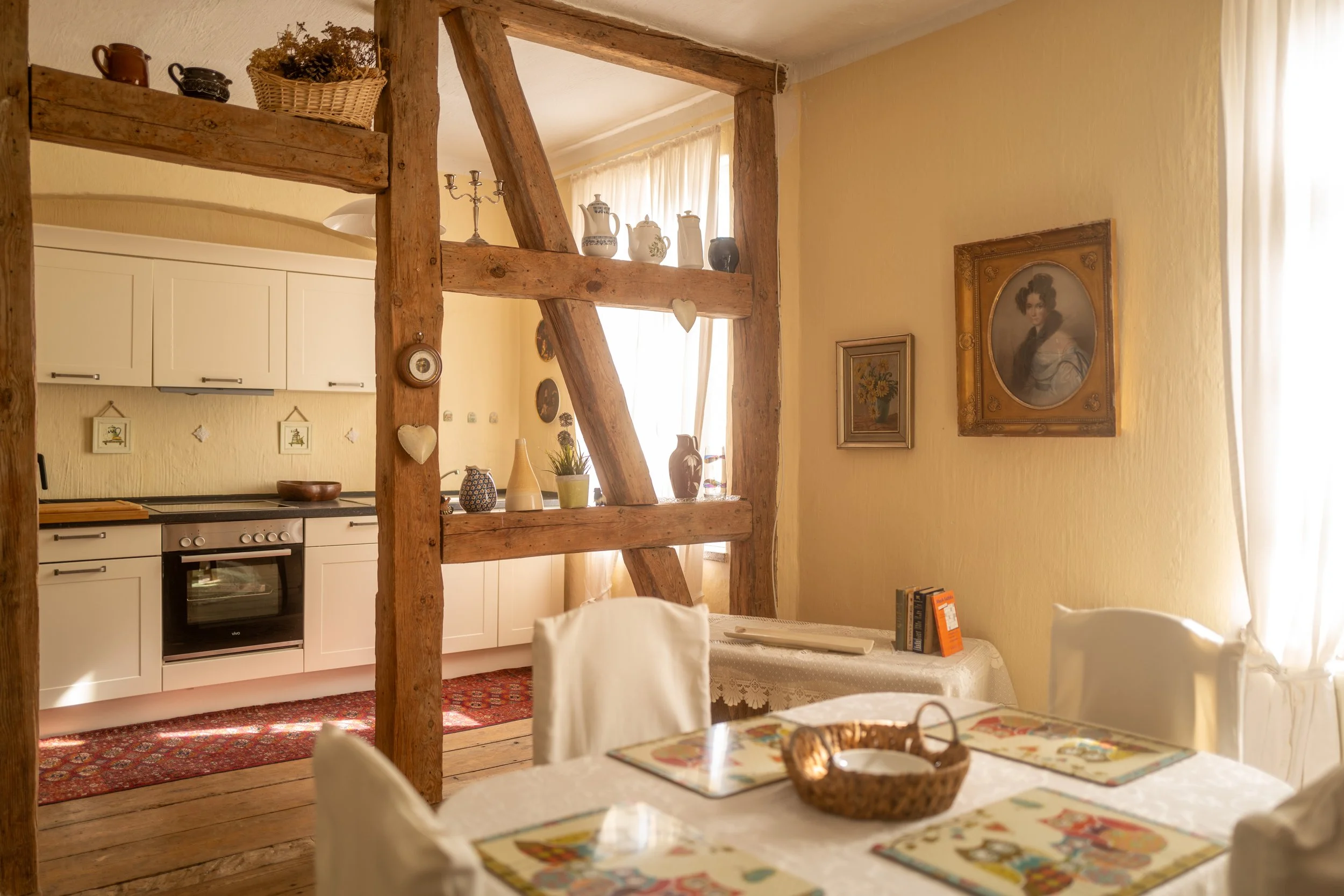 Ein Einrichtungsgegenstand in einer gemütlichen Küche, mit Kerzenständer, Töpfen, Vasen und gerahmten Bildern an der Wand, sowie einem Esstisch mit Tischdecken und Stühlen.