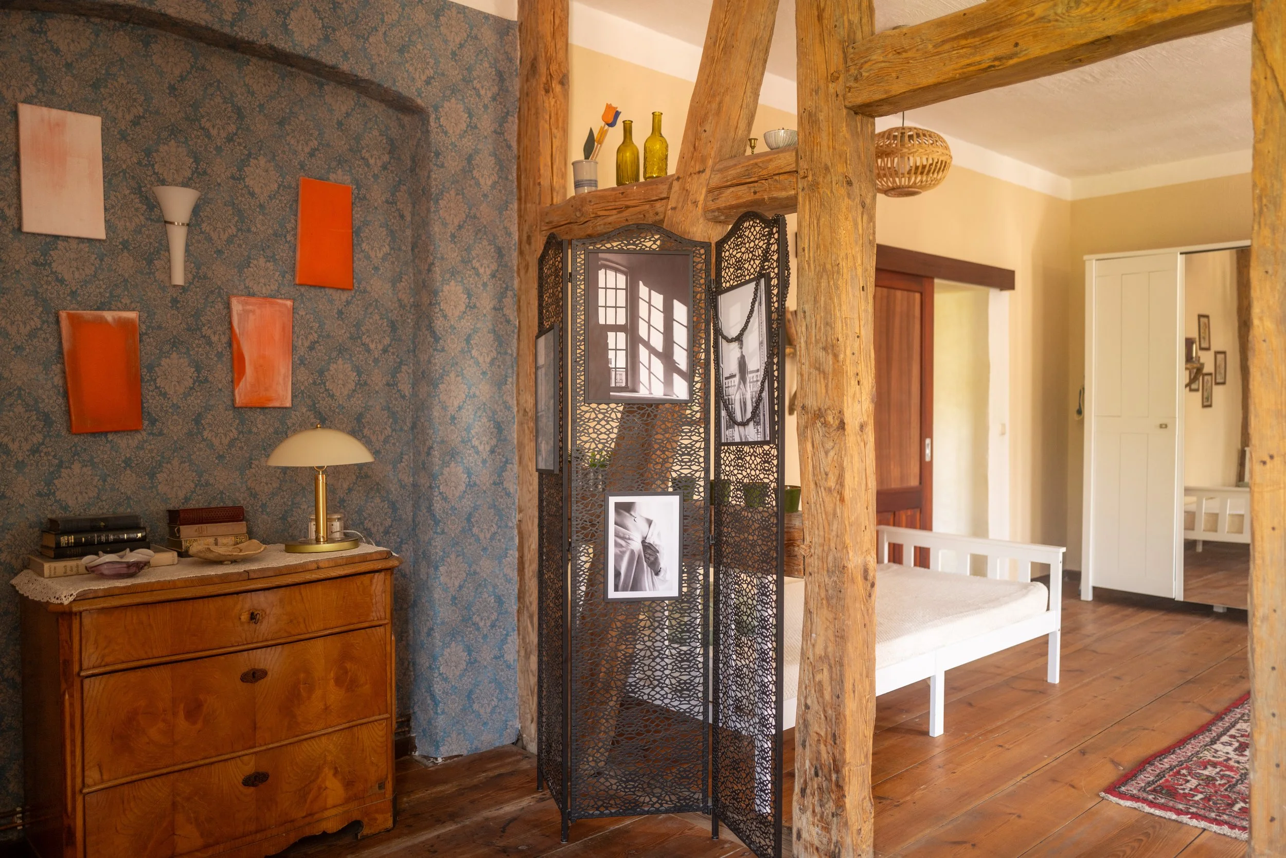 Innenansicht eines wohnlichen Raumes mit Holzdecke, -boden und -stützen, Wanddekor mit blauer Tapete, gemusterte Textilien, alten Büchern, Lampen, Vasen und Dekorationsgegenständen.