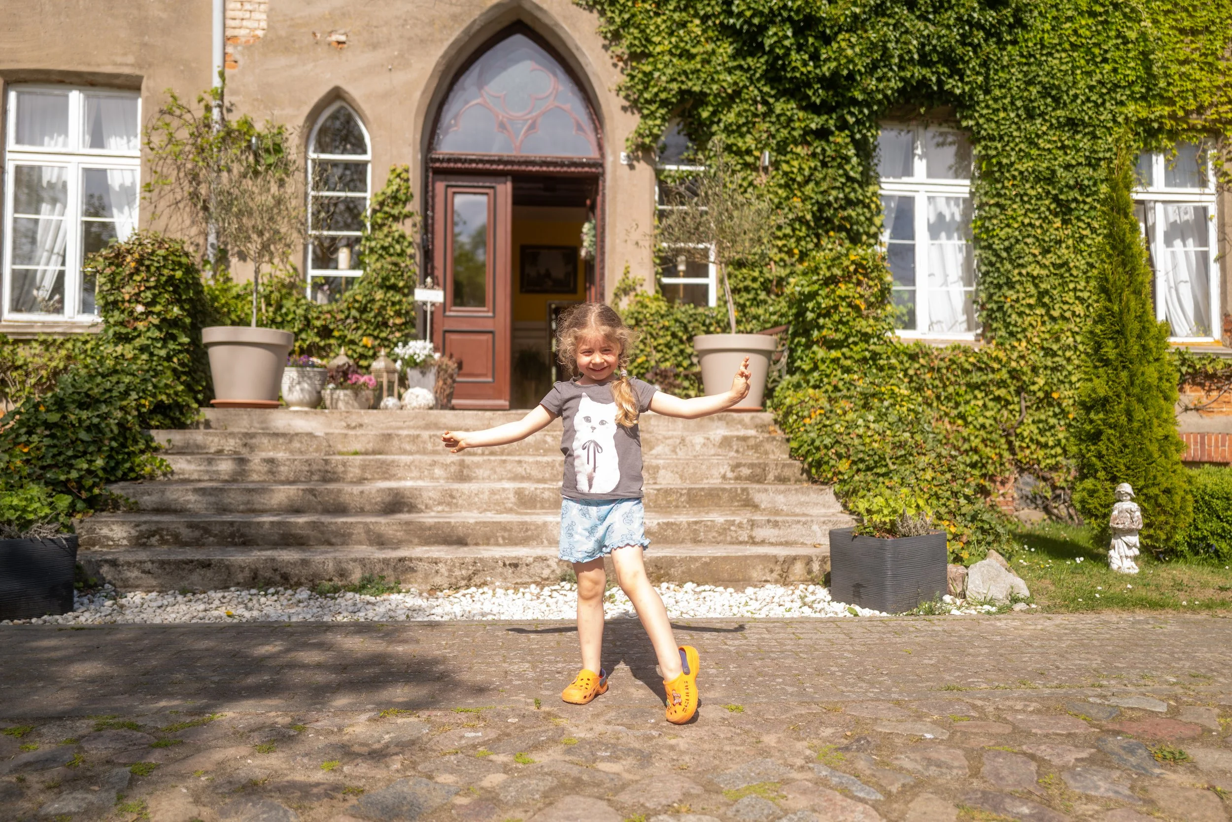 Ein lachendes junges Mädchen mit langen, lockigen Haaren, das auf einem gepflasterten Weg vor einem alten, bewachsenen Haus steht. Das Mädchen trägt ein graues T-Shirt mit einer Katzenillustration, hellblaue Shorts, orange Crocs und wirft die Arme fr