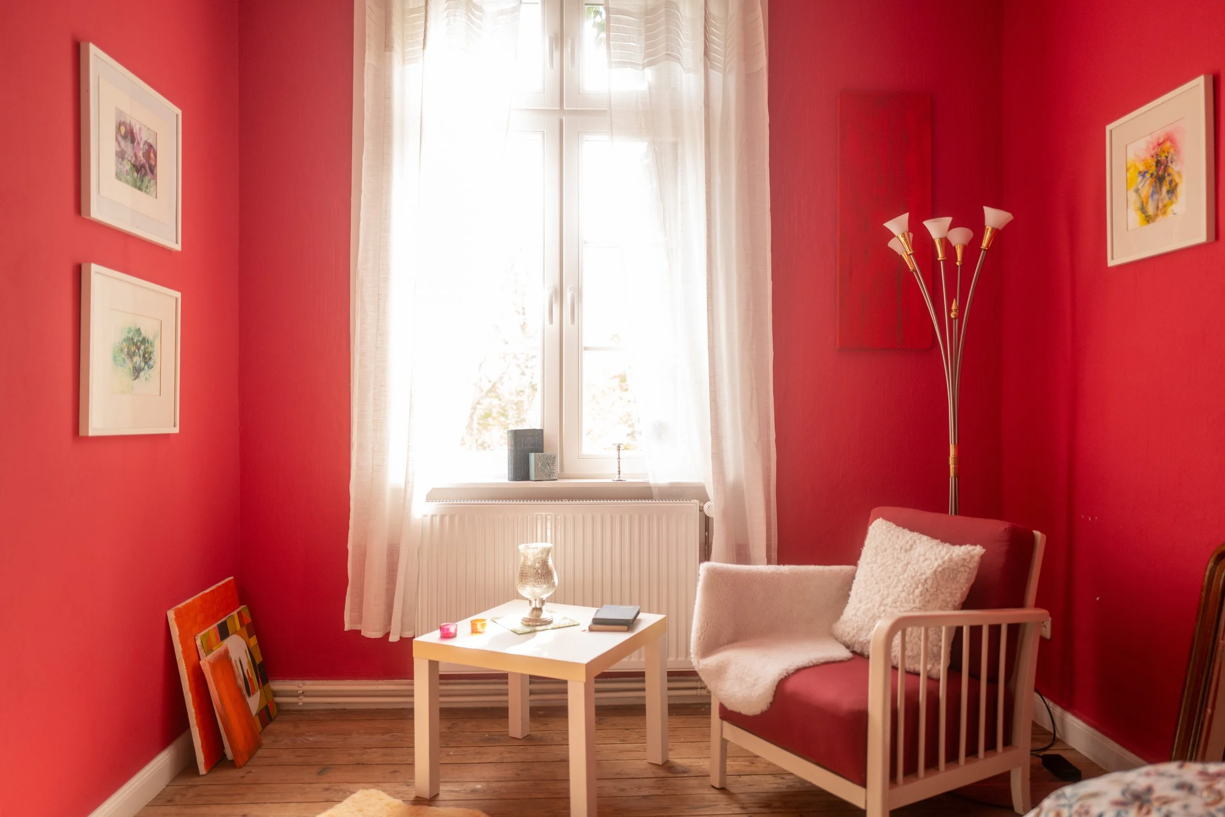 Ein gemütliches Wohnzimmer mit roten Wänden, weißen Vorhängen und mehreren Wandbildern. Es gibt einen weißen Sessel mit Kissen, einen kleinen weißen Tisch mit Dekorationen und eine Stehlampe.