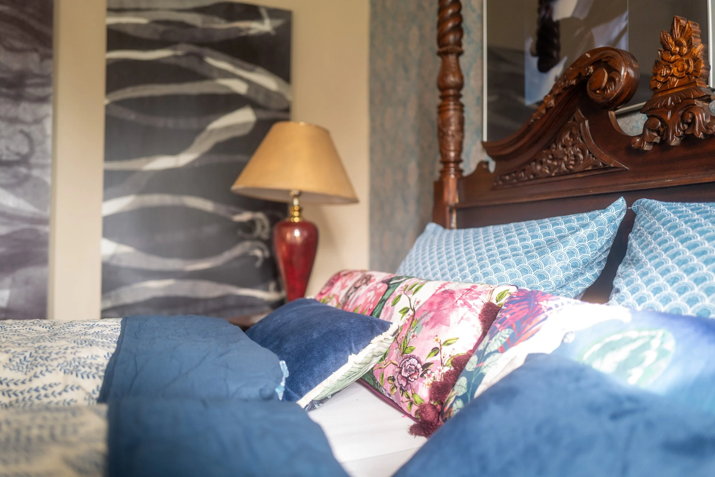 Bett mit vielfältigen bunten Kissen, darunter ein rosa Kissen mit floralem Muster, ein bläuliches Kissen mit Wellenmuster, und ein dunkelblaues Kissen.