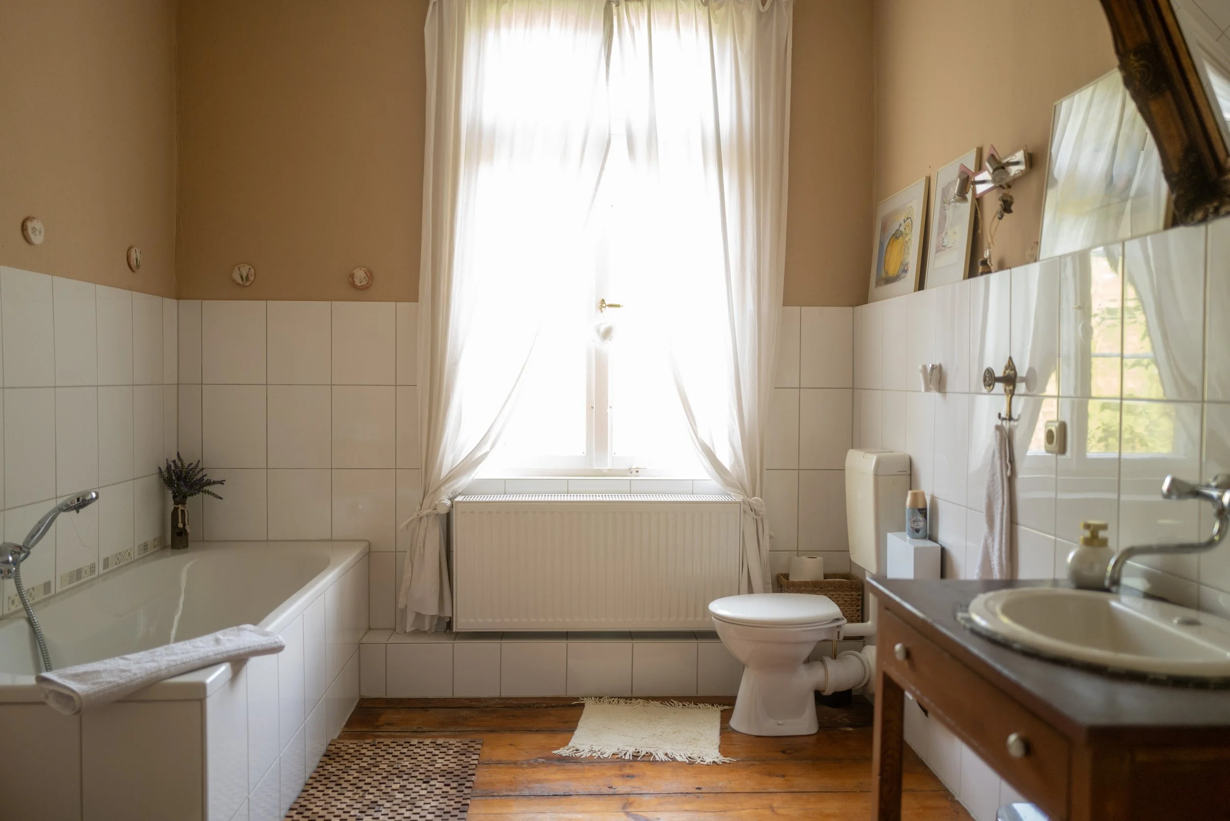 Ein helles Badezimmer mit weißen Fliesen an der Wand, einem Fenster mit weißen Vorhängen, einer Badewanne mit einem weißen Handtuch, einem Toilettenpapier und einem Waschtisch mit Spiegel und Dekorationen.