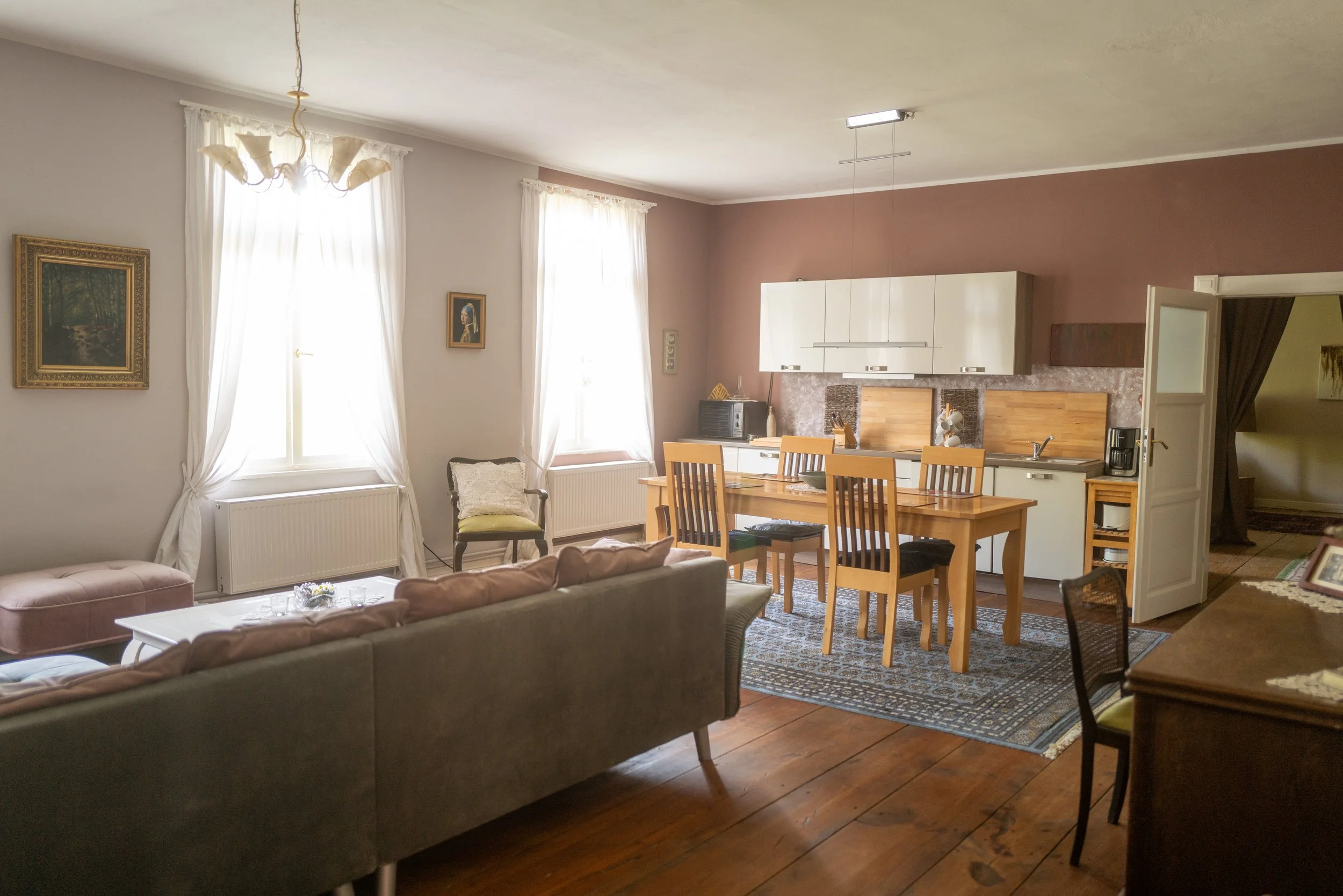 Ein Wohnzimmer mit weißen Vorhängen, einem Esstisch mit sechs Stühlen, einer Küche im Hintergrund und Wanddekorationen.