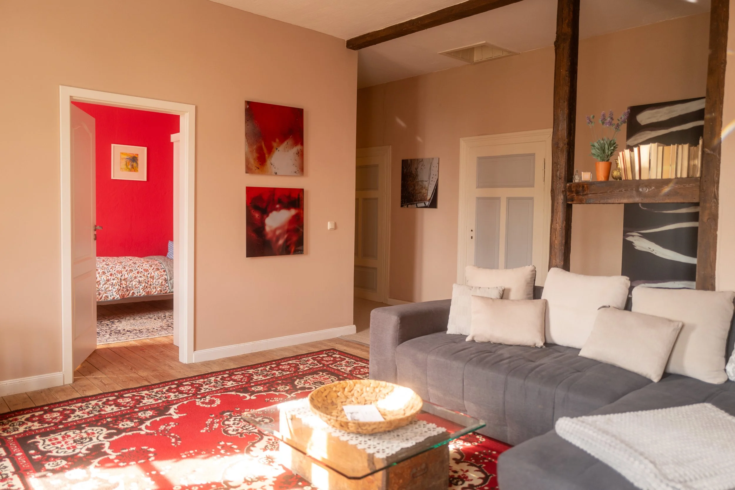 Ein Wohnzimmer mit grauem Sofa, weißen Kissen, einem Holzcouchtisch, rotem Teppich, Wandbilder, eine halb offene Tür zu einem Schlafzimmer mit roter Wand und buntem Bettbezug, und eine Regal mit Pflanzen und Büchern.