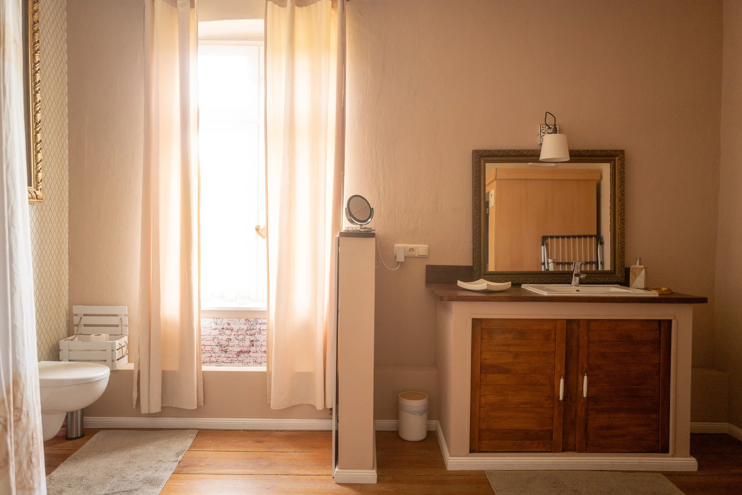 Ein Badezimmer mit einer Dusche, einem großen Fenster mit hellen Gardinen, einem Spiegel, einem Waschbecken, einem Holzschrank und weiteren Badezimmerutensilien.