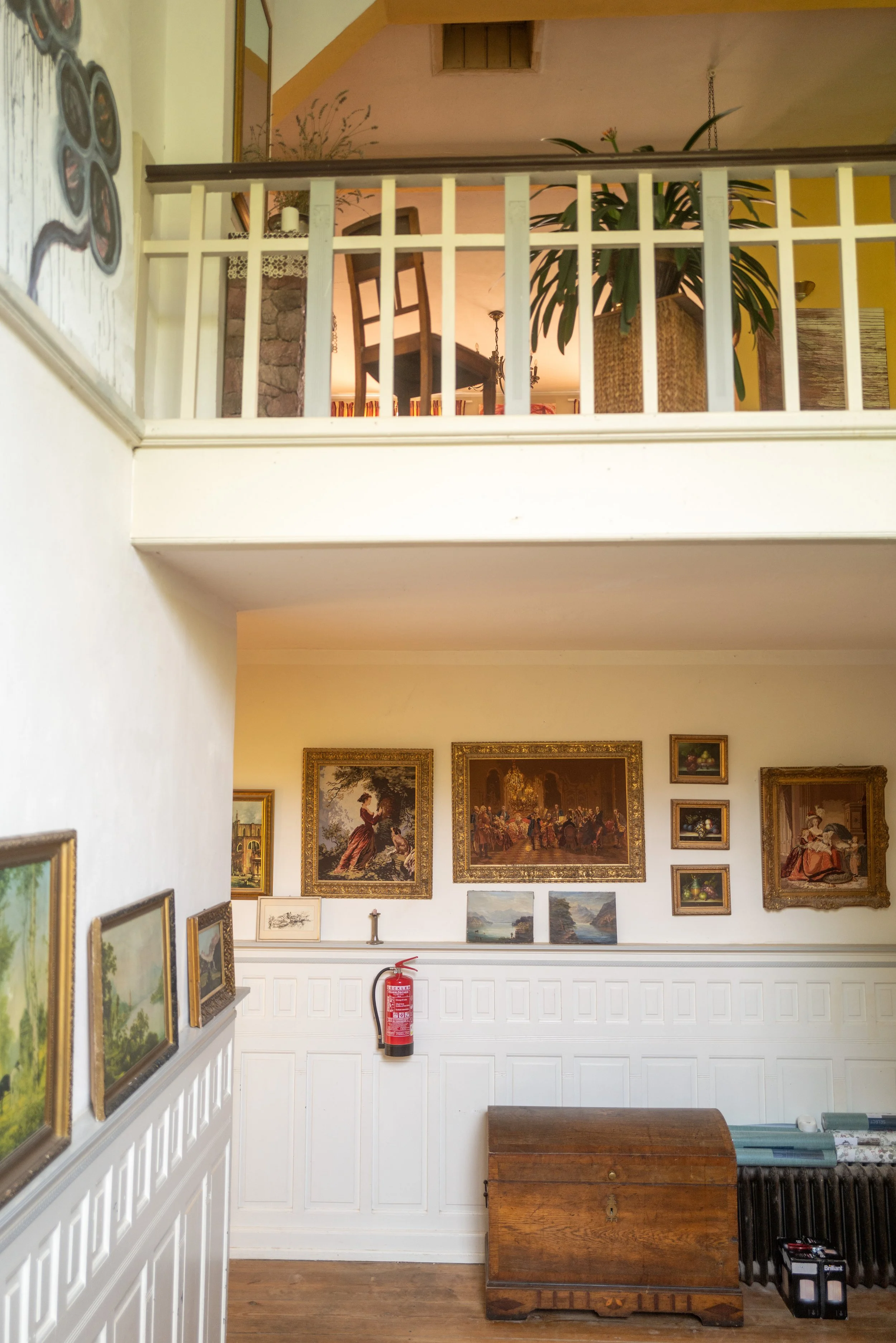 Blick in ein kunsthistorisches Museum mit einer Wand voller Gemälde und einem noch verschlossenen Holztruhe, auf dem Boden ein Feuerlöscher und eine Laterne.