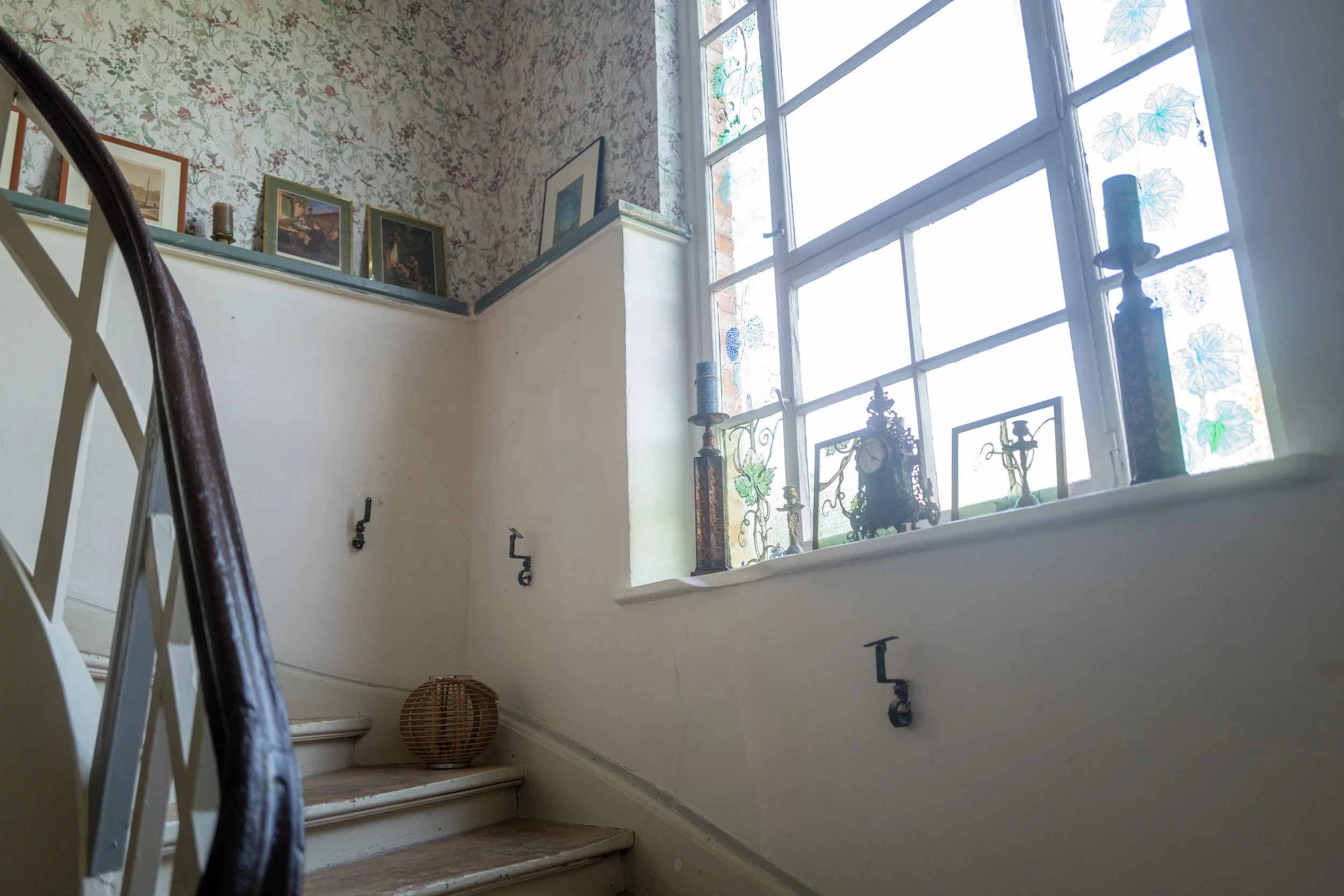 Innenansicht eines Hauses mit Treppe, Mann sitzt an einem Fenster, mit verschiedenen Dekorationsgegenständen und Bildern an der Wand.