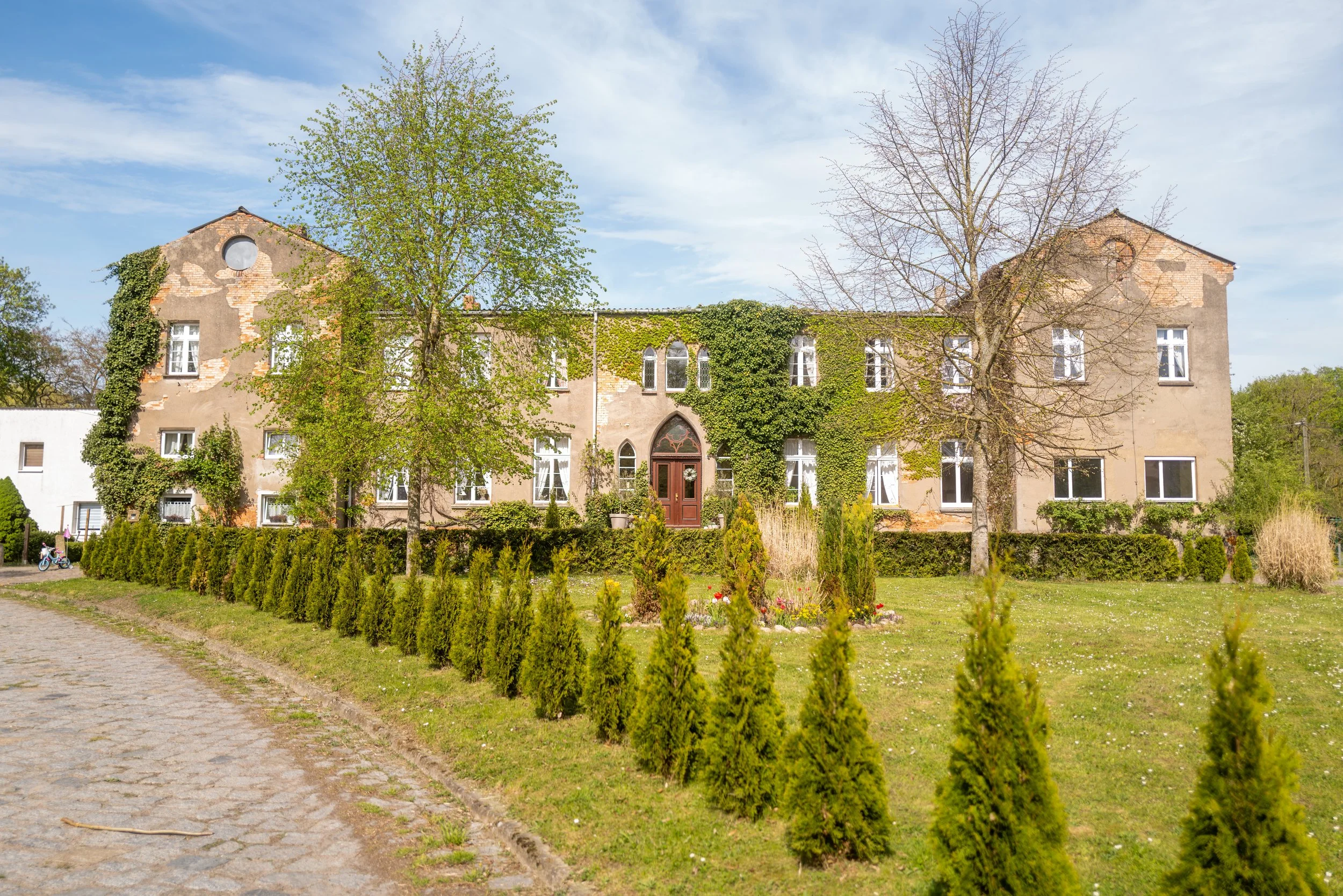 Ein altes, leicht verfallenes Haus mit mehreren Fenstern, teilweise von Efeu überwuchert, umgeben von Bäumen und einem Garten, bei blauem Himmel.