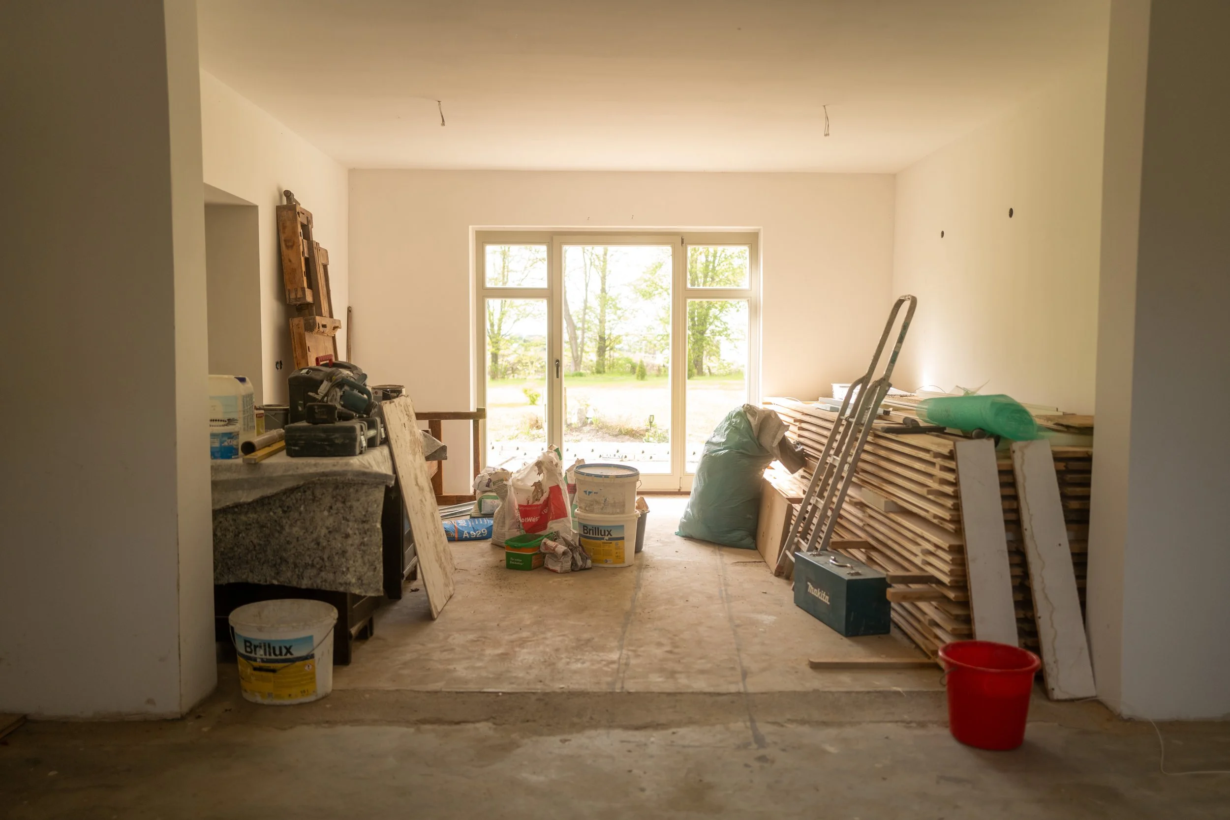Innenansicht eines Raums im Bau oder Renovierungsprozess, mit Baumaterialien, Werkzeugen und unvollendeter Wand. Große Fenster lassen Tageslicht herein und bieten Blick auf einen grünen, bewaldeten Außenbereich.