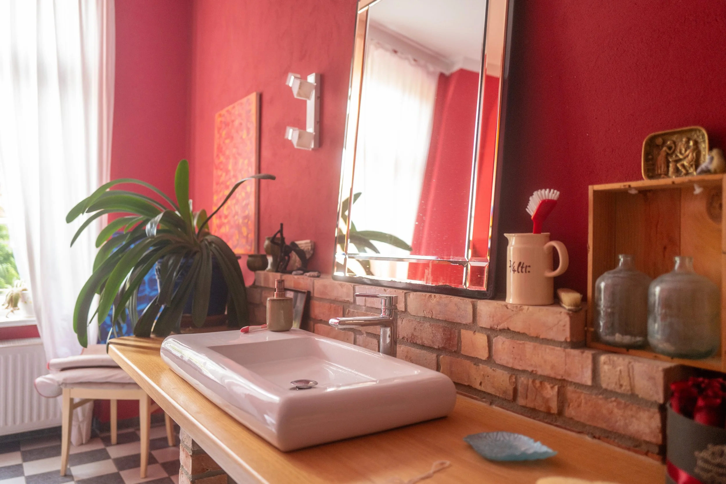 Ein Badezimmer mit roter Wand hinter einem weißen Waschbecken und einem großen Spiegel. Es gibt eine braune Holzablage mit verschiedenen Gegenständen wie einem Keramikbecher, Flaschen, und Dekorationsgegenständen. Ein grüner Pflanzenzweig ist im Vordergrund.