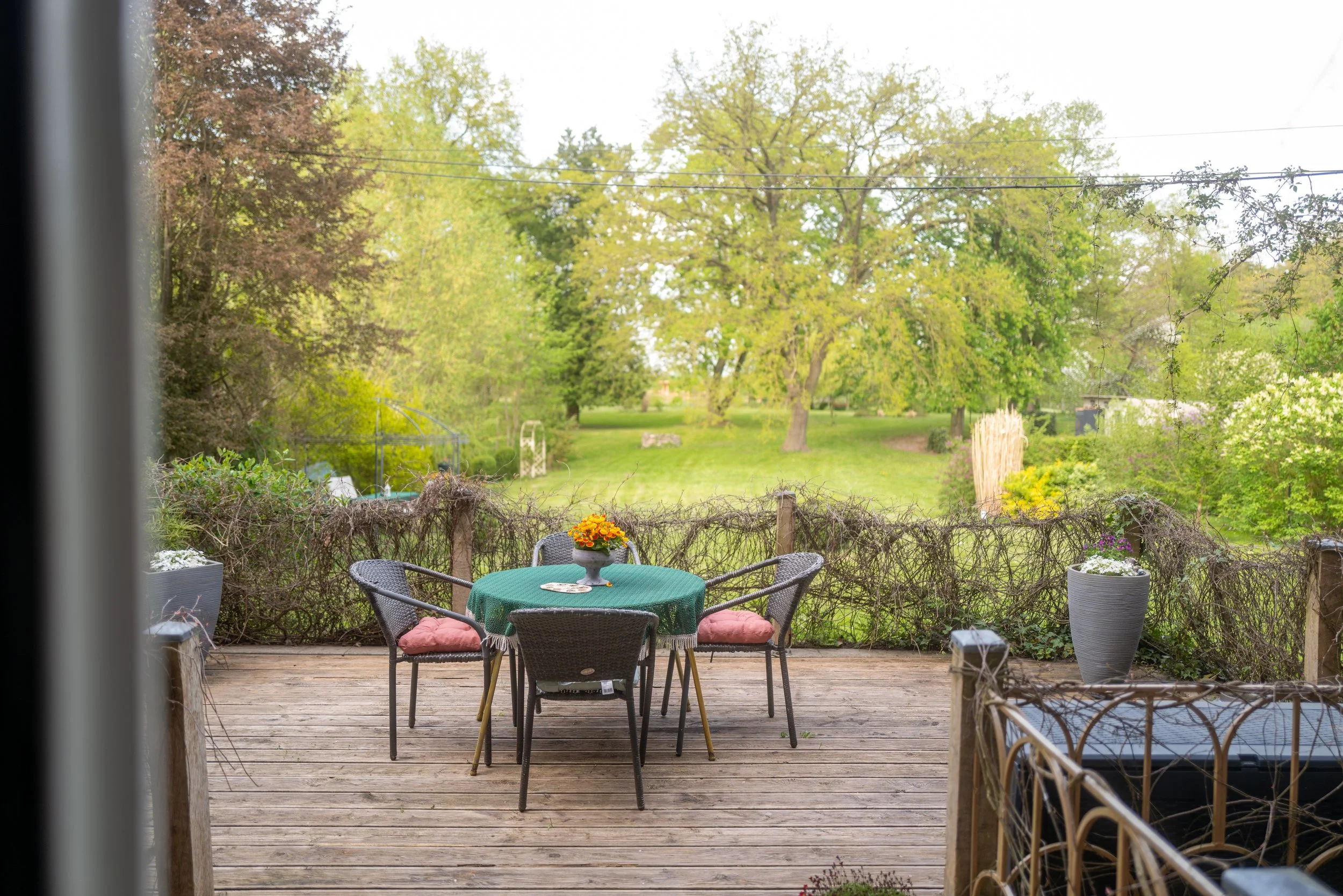 Blick auf einen grünen Garten mit Bäumen durch eine offene Terrassentür, Gartenmöbel und bepflanzte Töpfe auf einer Holzterrasse im Vordergrund.