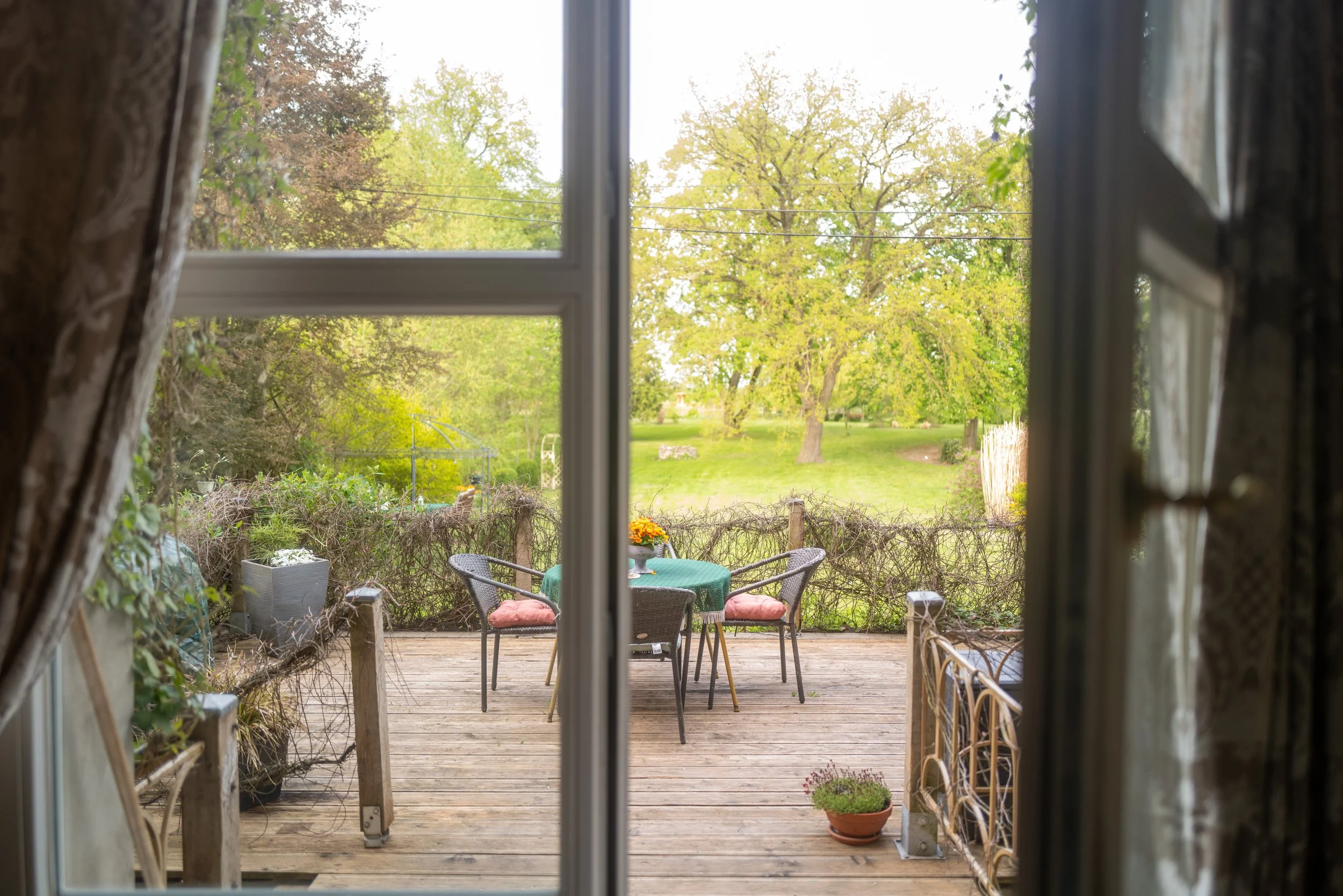 Blick durch eine offene Fenstertür auf eine Terrasse mit einem runden Tisch und vier Stühlen, umgeben von Pflanzen und einem Garten mit grünen Bäumen.