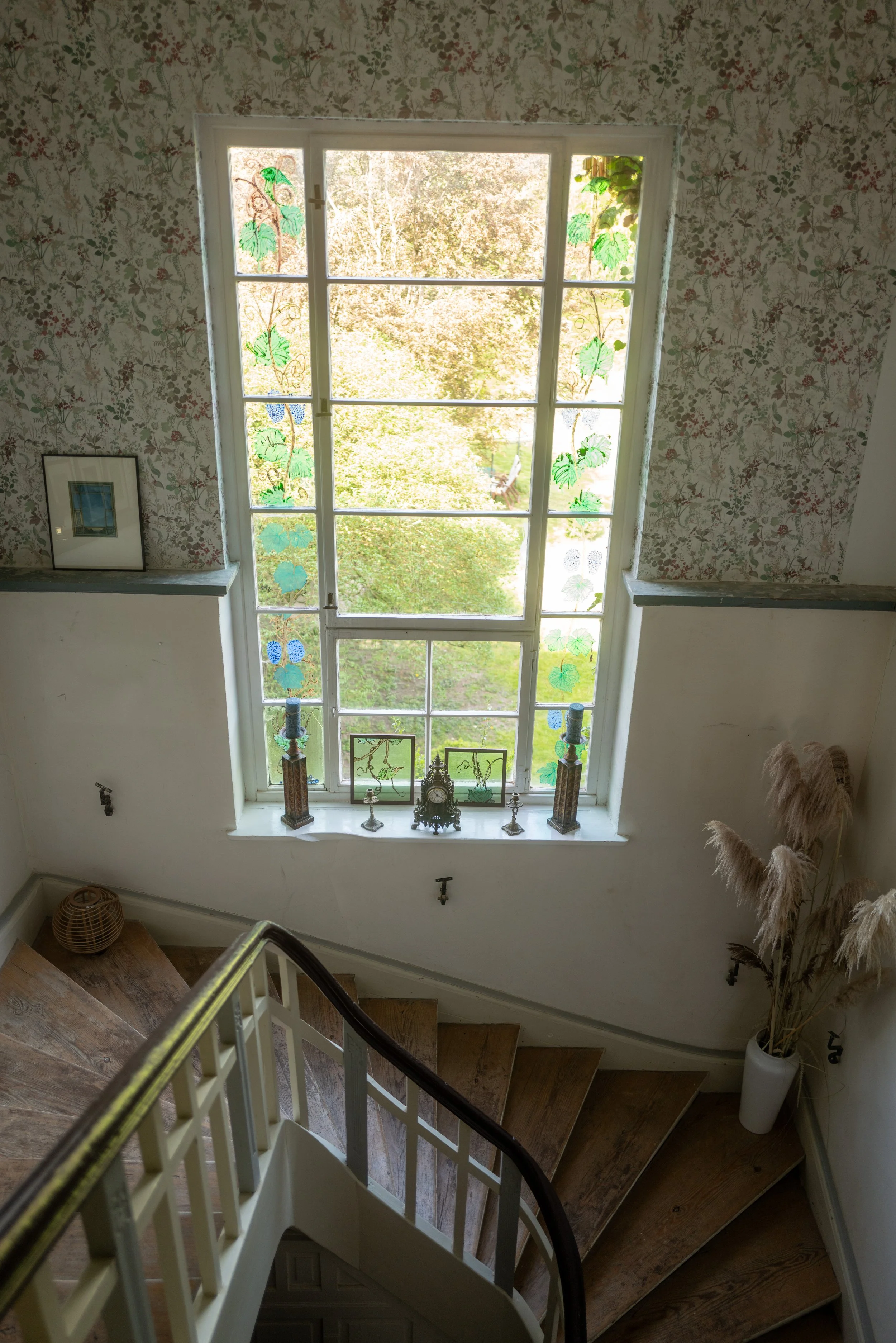 Innenseite eines Hauses mit einem großen Fenster, das grüne und blaue dekorative Lamellen mit Weinrebenmuster hat. Es gibt eine Treppe mit Holzstufen und eine weiße Geländer, unter dem Fenster sind allerlei Dekorationsgegenstände platziert, darunter 
