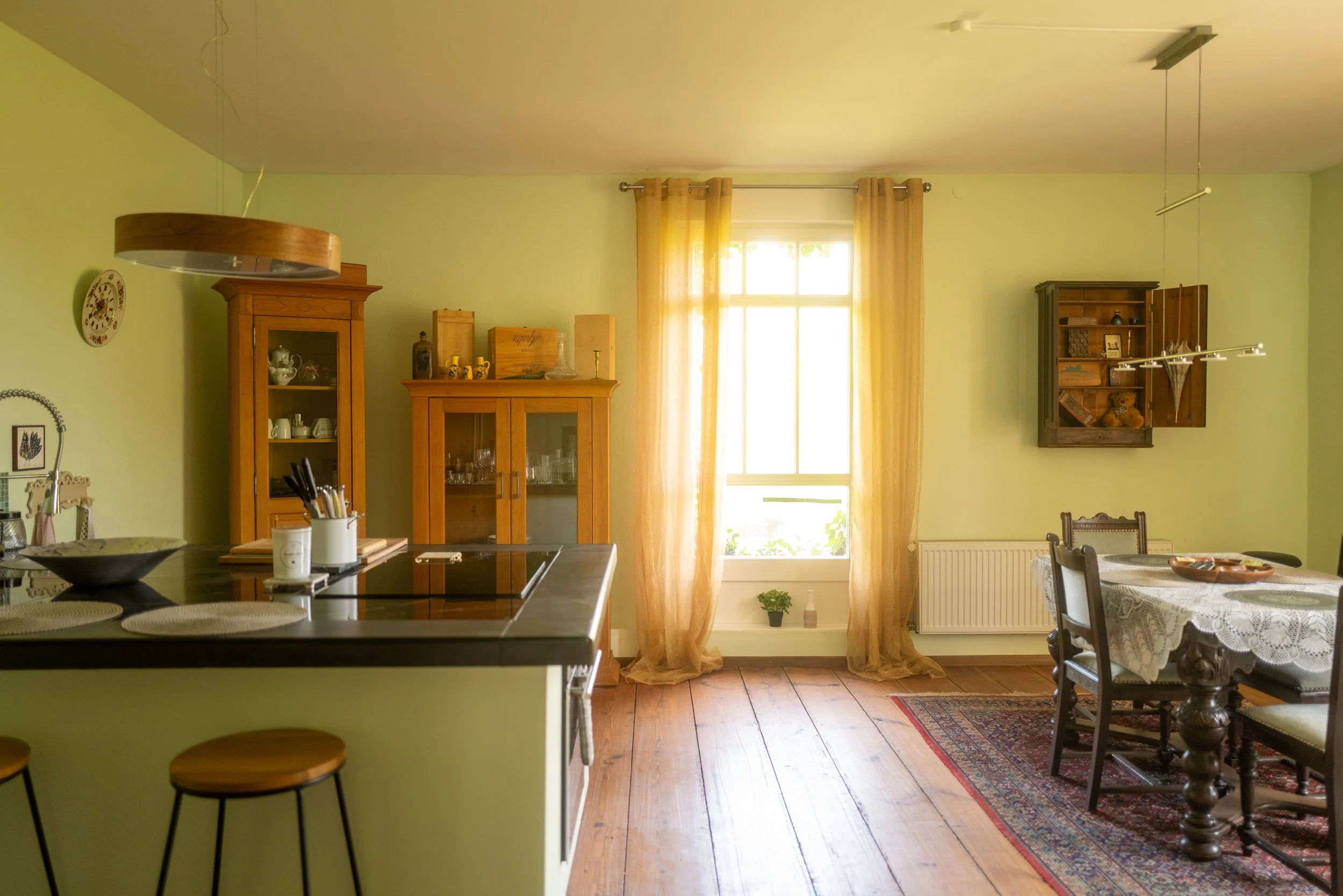 Einwohnküche mit grün gestrichenen Wänden, Holzmöbeln, einem Esstisch mit weißen Tischdecken, gelben Gardinen vor einem Fenster, Holzboden und einem dunklen Teppich.