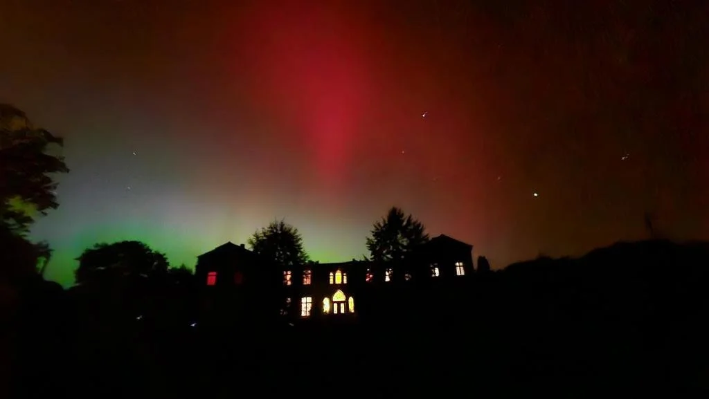 Ein dunkles Haus mit beleuchteten Fenstern vor einem Himmel mit Nordlichtern und Sternen.
