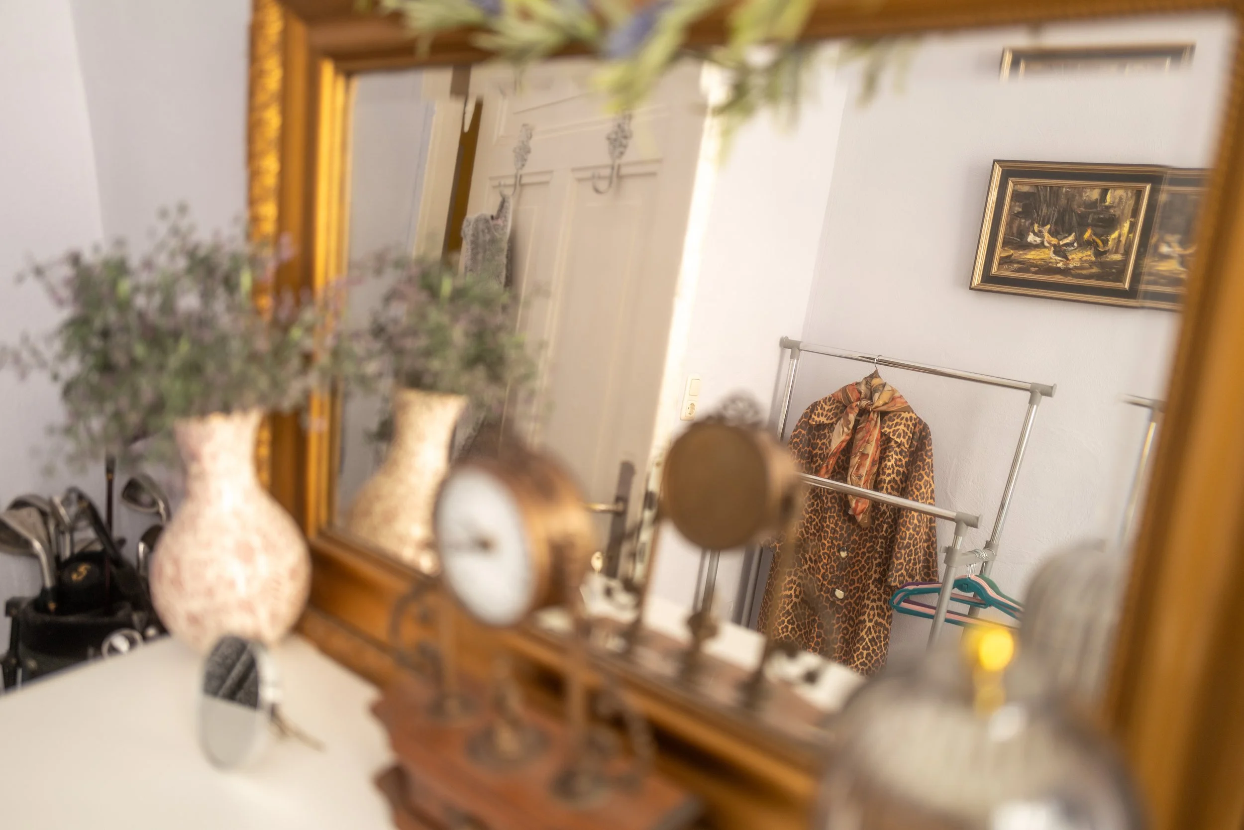 Ein spiegelbildlicher Blick in einen Flur mit einem Wandbild und Kleidung auf einer Stange, im Vordergrund eine Dekoration aus Vasen, Pflanzen und Schuhen, die Bilder sind unscharf, das Hauptbild zeigt eine Tür, einen Rahmen und eine Garderobe.