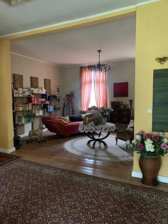 Ein Wohnzimmer mit gelben Wänden, einem roten Sofa, einem Couchtisch mit einer platzierten Vasen mit Blumen, einem Teppich, einem Bücherregal, einer Wanddekoration, einem Fenster mit roten und weißen Vorhängen und einer Deckenlampe.