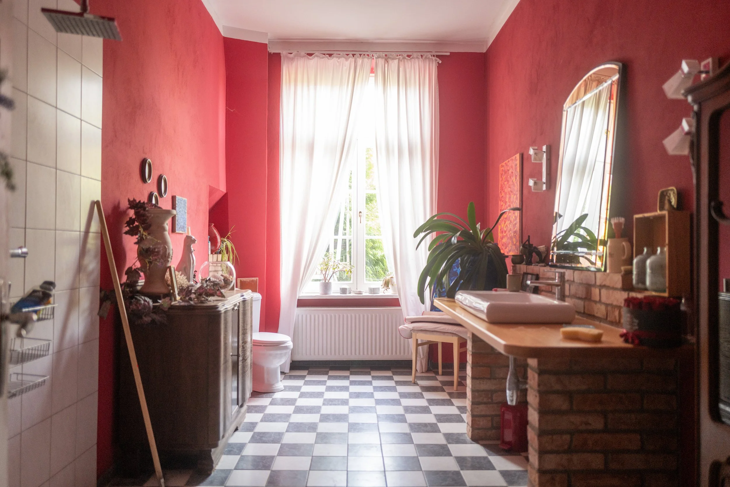 Ein Raum mit roten Wänden, einer großen Fensterfront mit weißen Vorhängen, mehreren Pflanzen, einem Spiegel, einer Holzbank und weiteren Einrichtungsgegenständen.