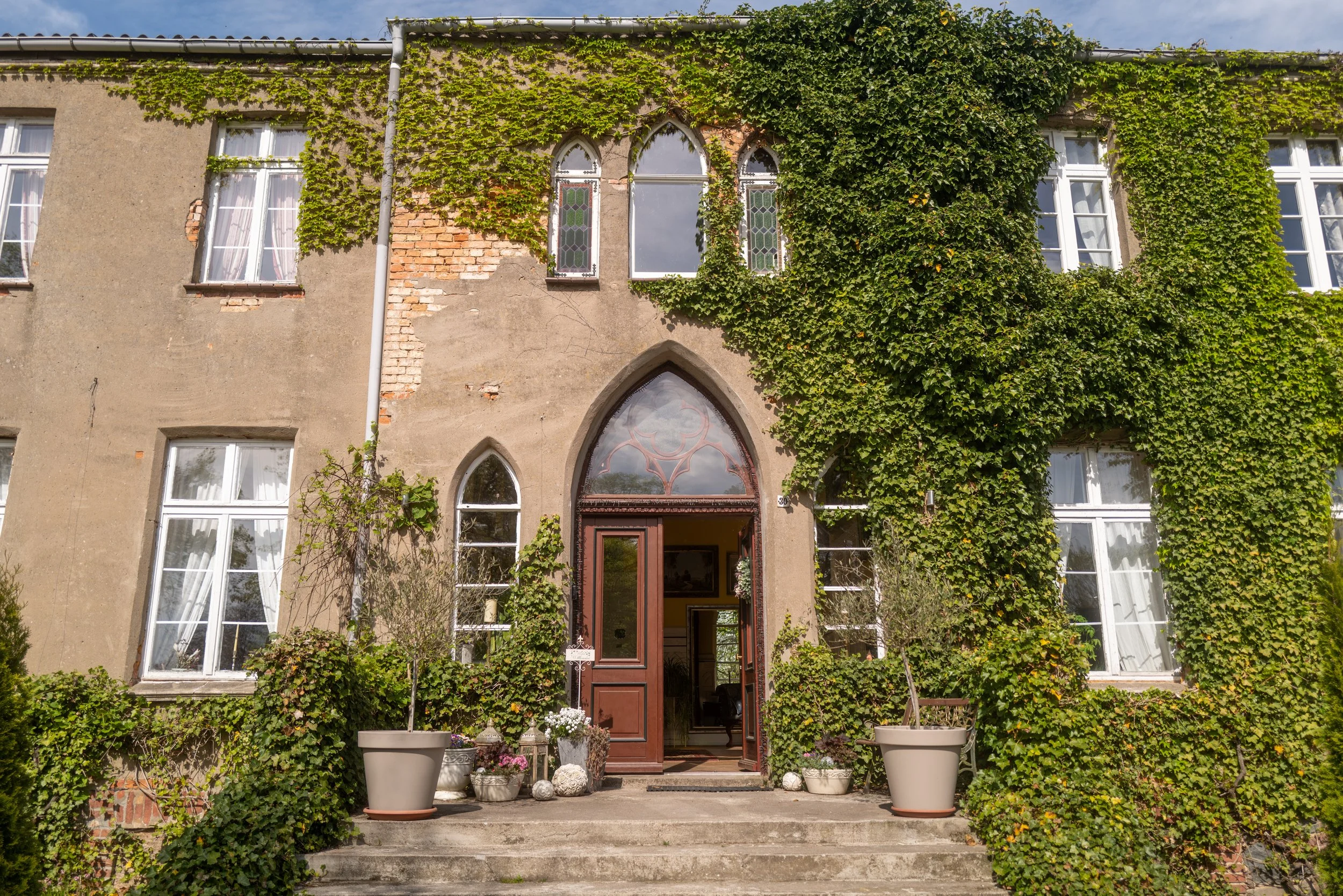 Altbau mit Graufasuren, teilweise verputzt, mit mehreren Fenstern und viel Efeu an der Fassade. Im Eingangsbereich befinden sich verschiedene Töpfe mit Pflanzen und Steine.