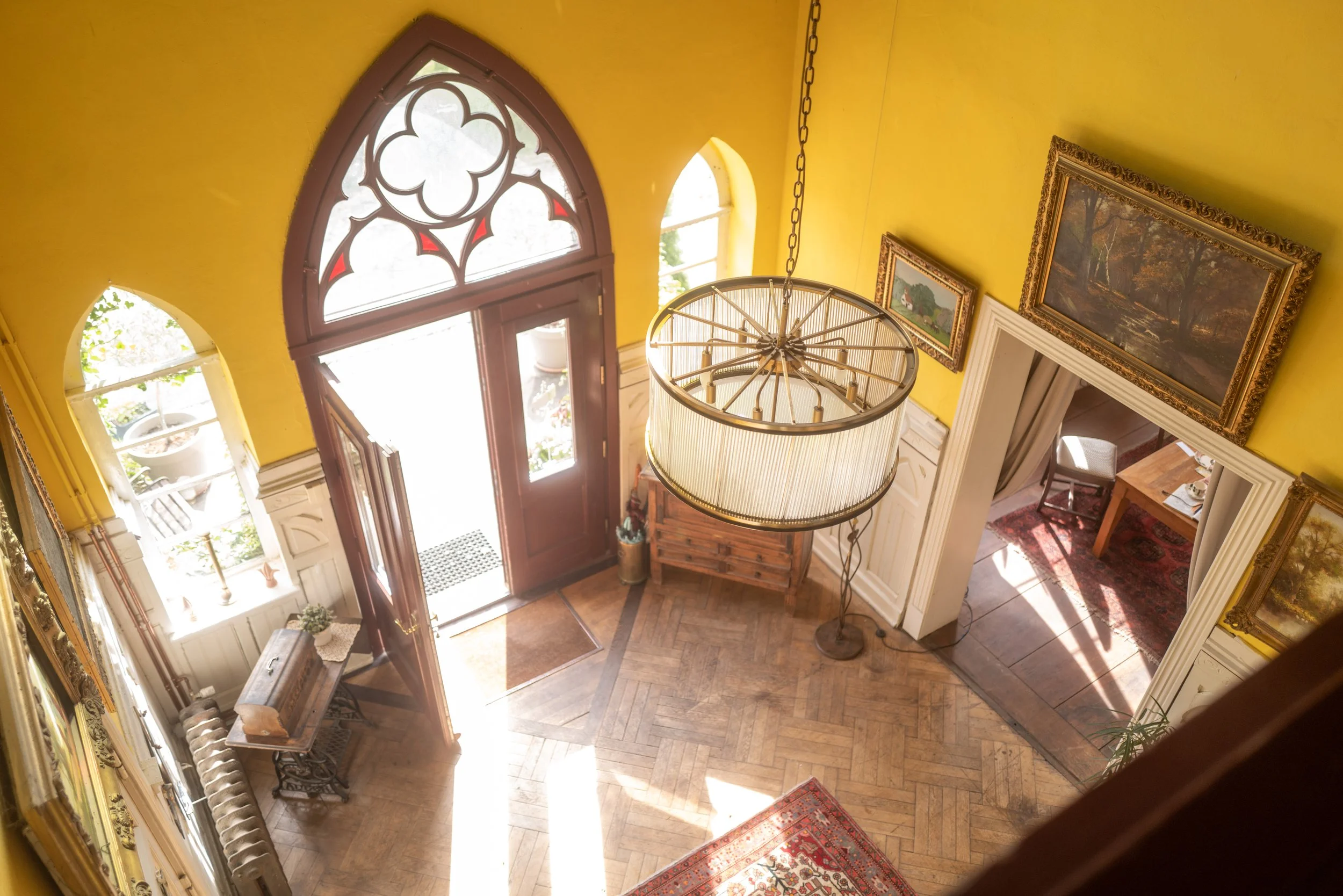 Blick in eine elegant eingerichtete Lobby mit gelben Wänden, einem prunkvollen Kronleuchter, Gemälden in goldenen Rahmen und einem Holztisch vor der Tür, die ins Freie führt.