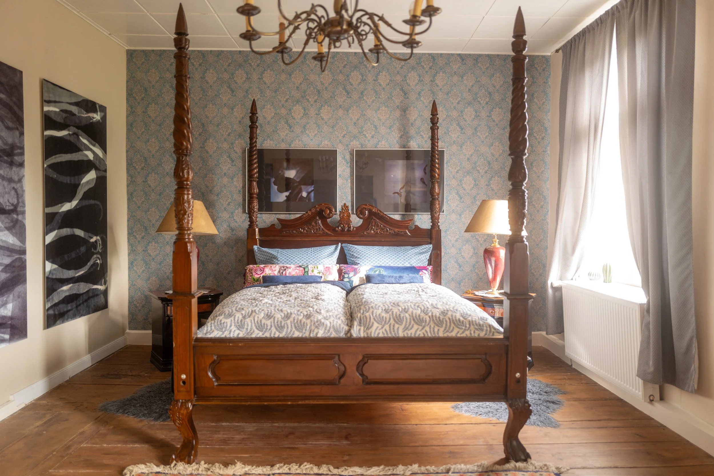 Ein Holz-Karussellbett in einem Schlafzimmer, mit vier Posten und einem geschnitzten Kopfteil, vor einer Wand mit blumigem Tapetenmuster, flankiert von zwei Nachttischen mit Lampen, und einem Fenster mit weißen Vorhängen auf der rechten Seite.