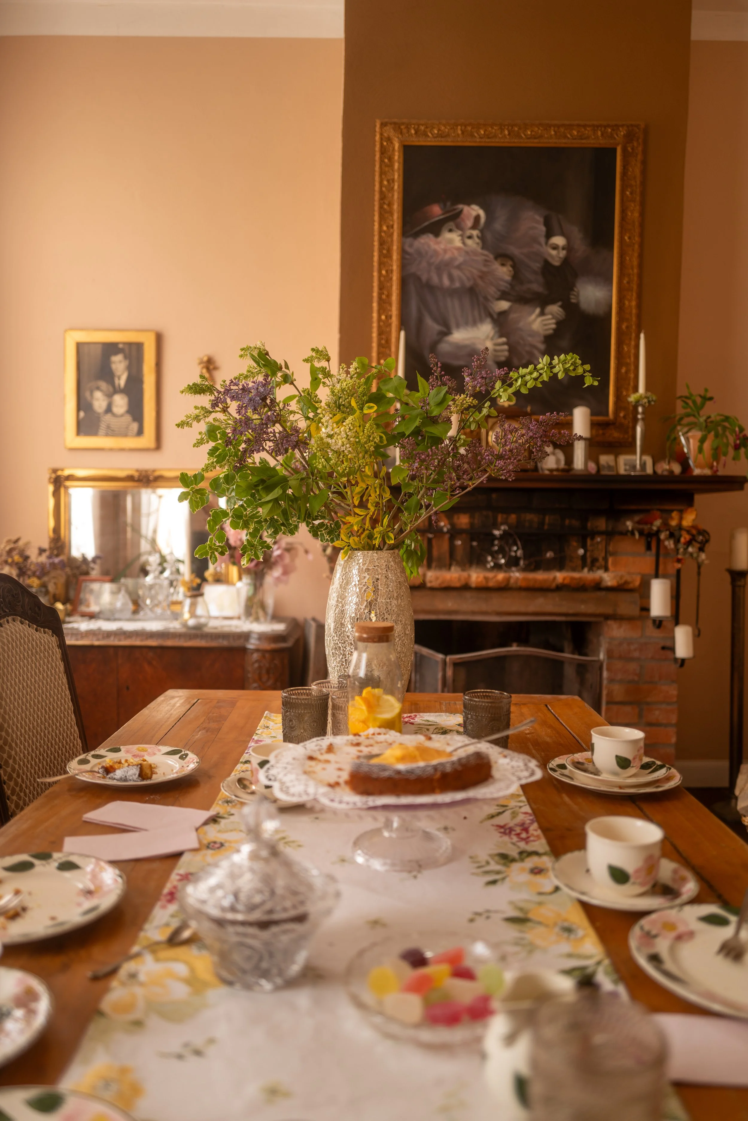 Ein festlich gedeckter Esstisch mit Kuchen, Tassen, Gläsern und einem Blumenstrauß im Vordergrund in einem gemütlichen Raum, mit Bildern an der Wand und Kerzen auf dem Kamin im Hintergrund.