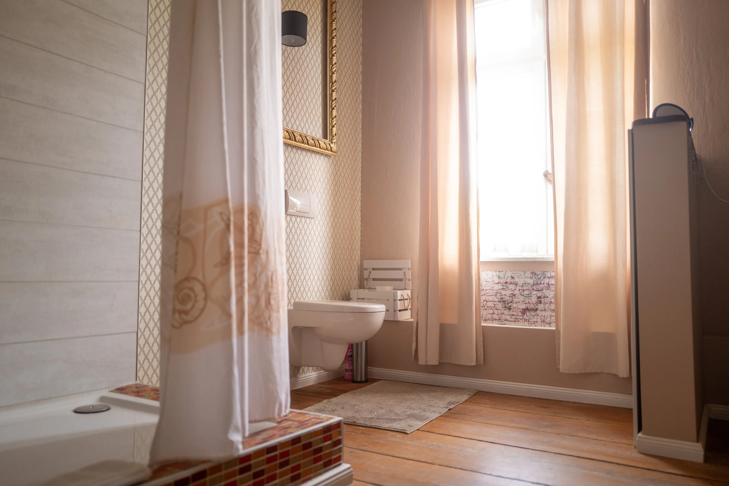Ein Badezimmer mit einer Toilette, einem Waschtisch, einem großen Fenster mit Vorhängen, einem Teppich auf dem Boden und einem großen Spiegel an der Wand.