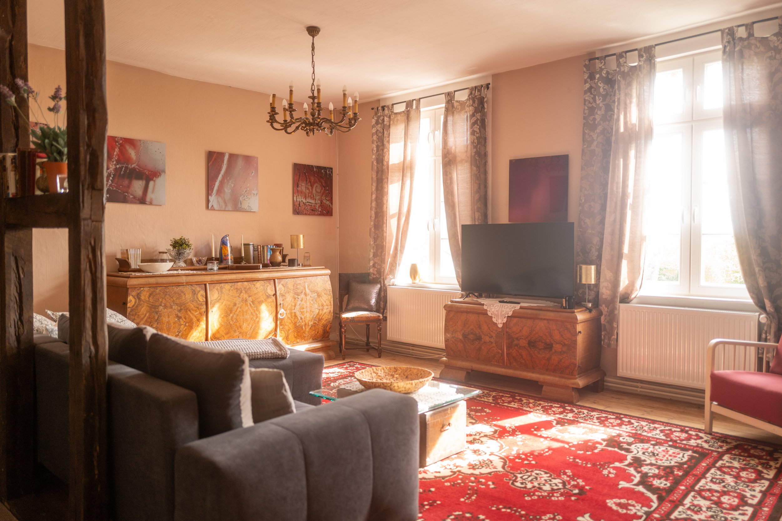 Wohnzimmer mit Sofa, Holzsideboard, Fernseher, große Fenster mit Gardinen, Kronleuchter, Teppich, Bild auf Wand, Wohnzimmerdeko, Kinderbett im Hintergrund.