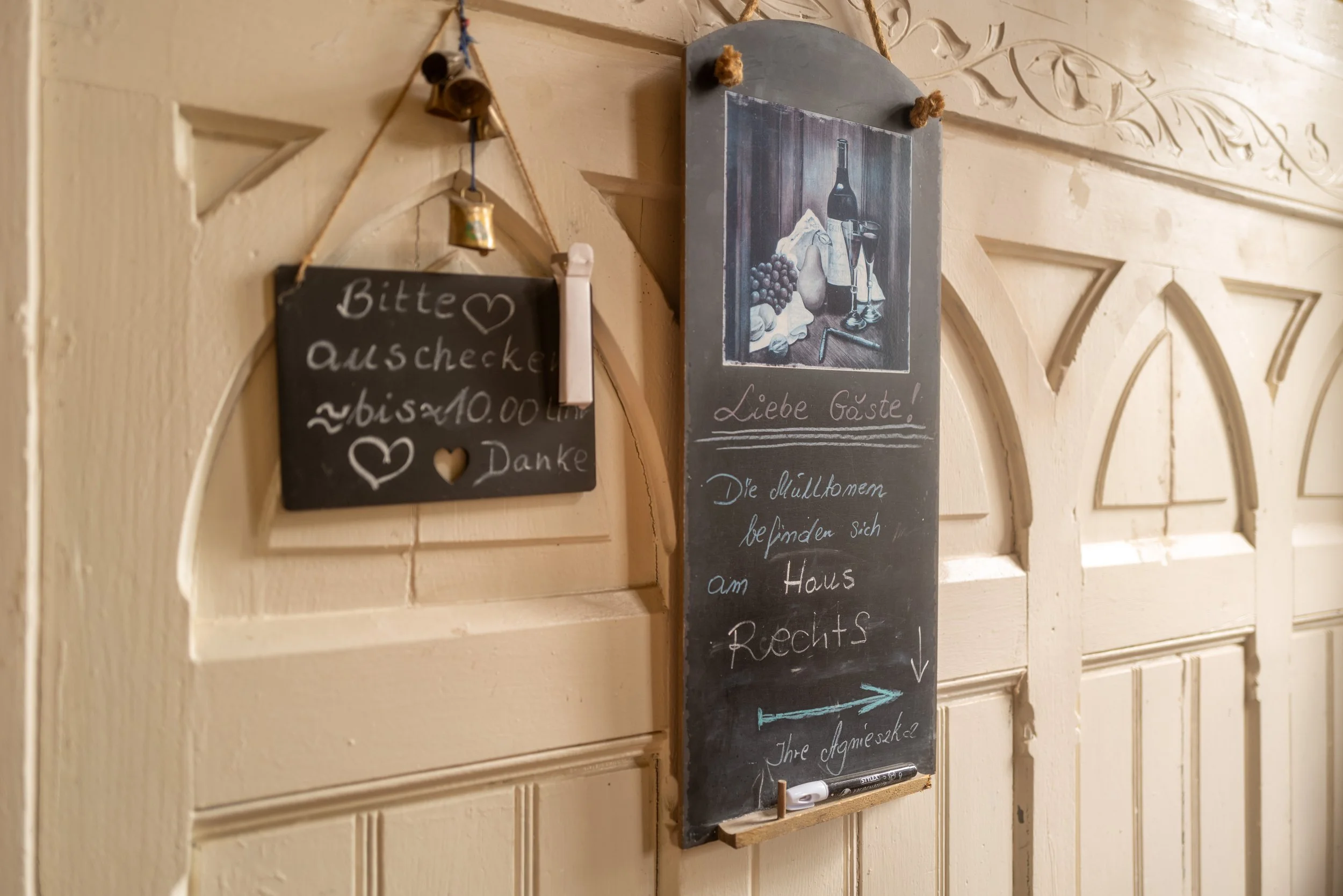 Ein Wandtafelschild mit handgeschriebenem Text in einer klassischen deutschen Küche. Das Schild begrüßt Gäste und erklärt, dass die Familie sich im Haus befindet. Es ist auf einer holzverzierten Wand angebracht.