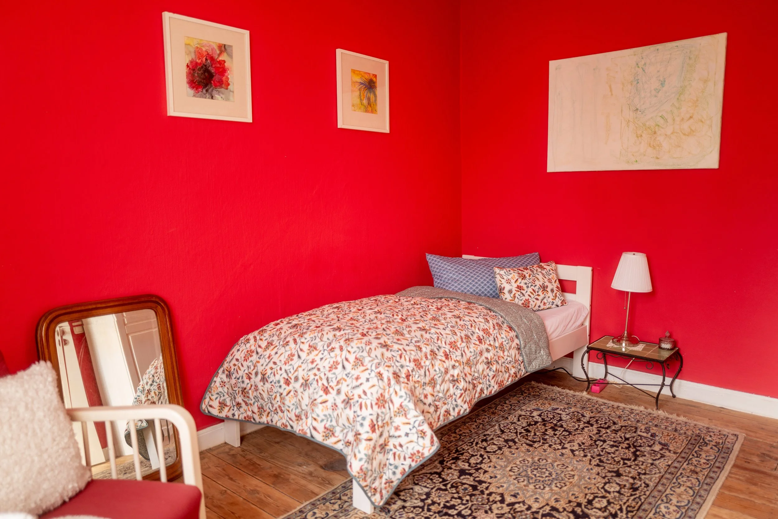 Ein Schlafzimmer mit roter Wand, einem weißen Bett mit buntem Bettbezug, drei Kissen, einem kleinen Nachttisch mit einer Lampe, einem Teppich und Wandbildern.