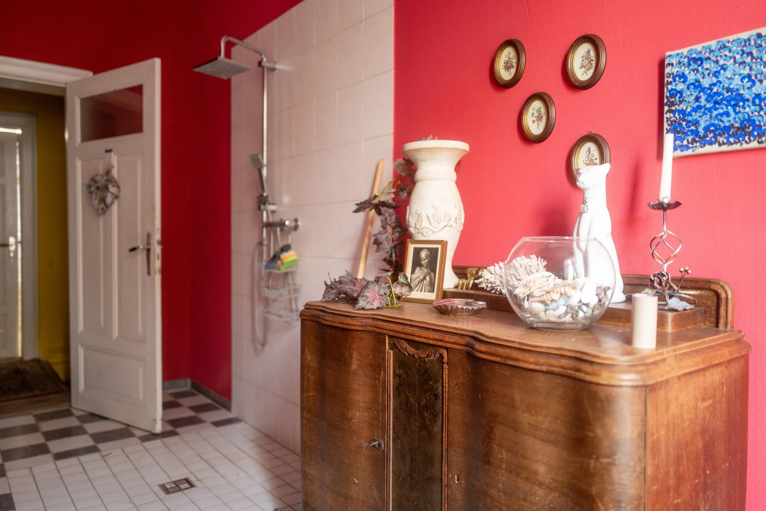 Ein Holzschrank vor einer roten Wand, dekoriert mit Vasen, Bildern und Kerzen, im Hintergrund eine Toilette und eine Dusche.