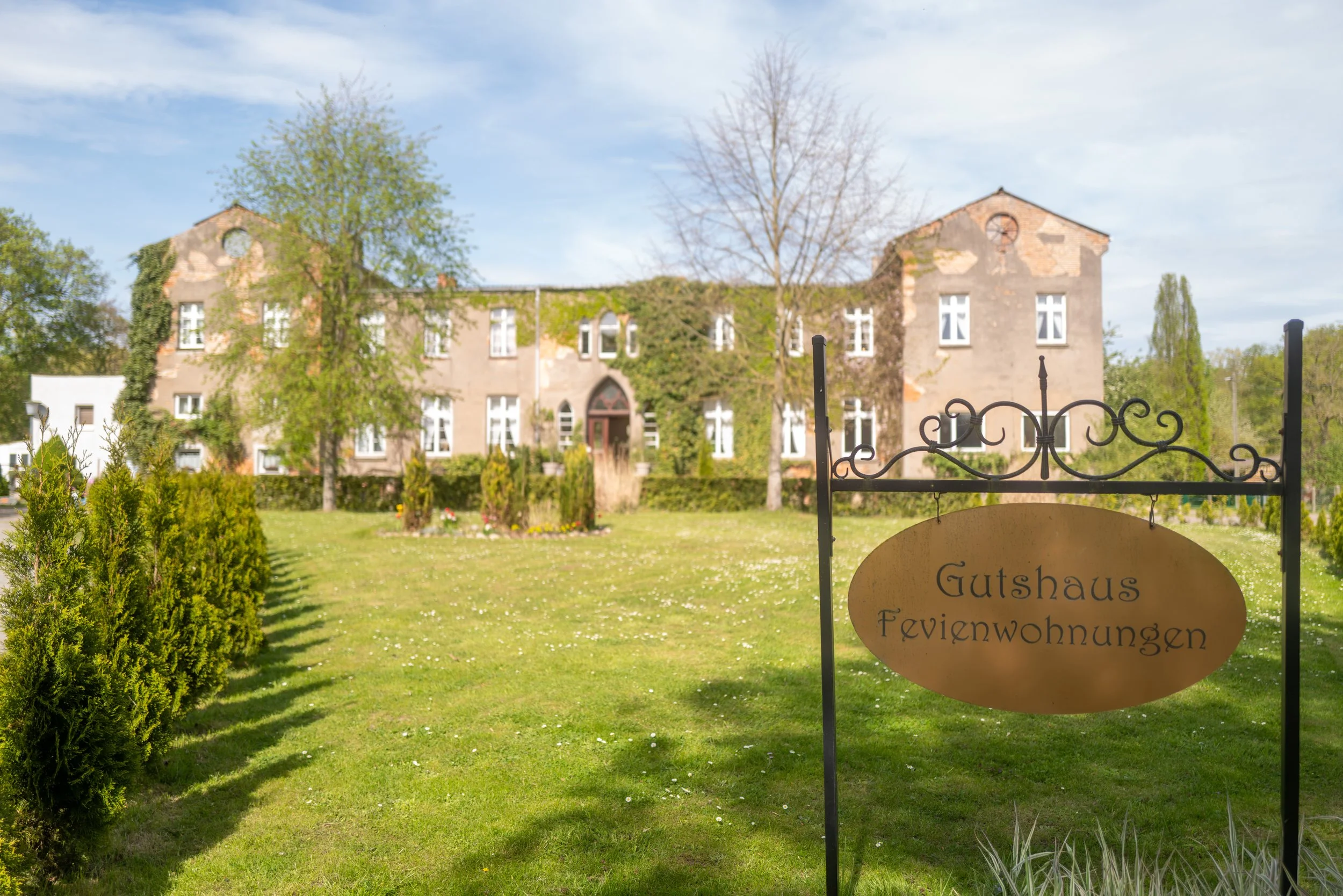 Ein Herrenhaus mit einer Schlossmauer im Hintergrund, umgeben von Bäumen und einem gepflegten Garten. Am Tor ist eine Schilder mit der Aufschrift 'Gutshaus Feyenwohnngen' befestigt.