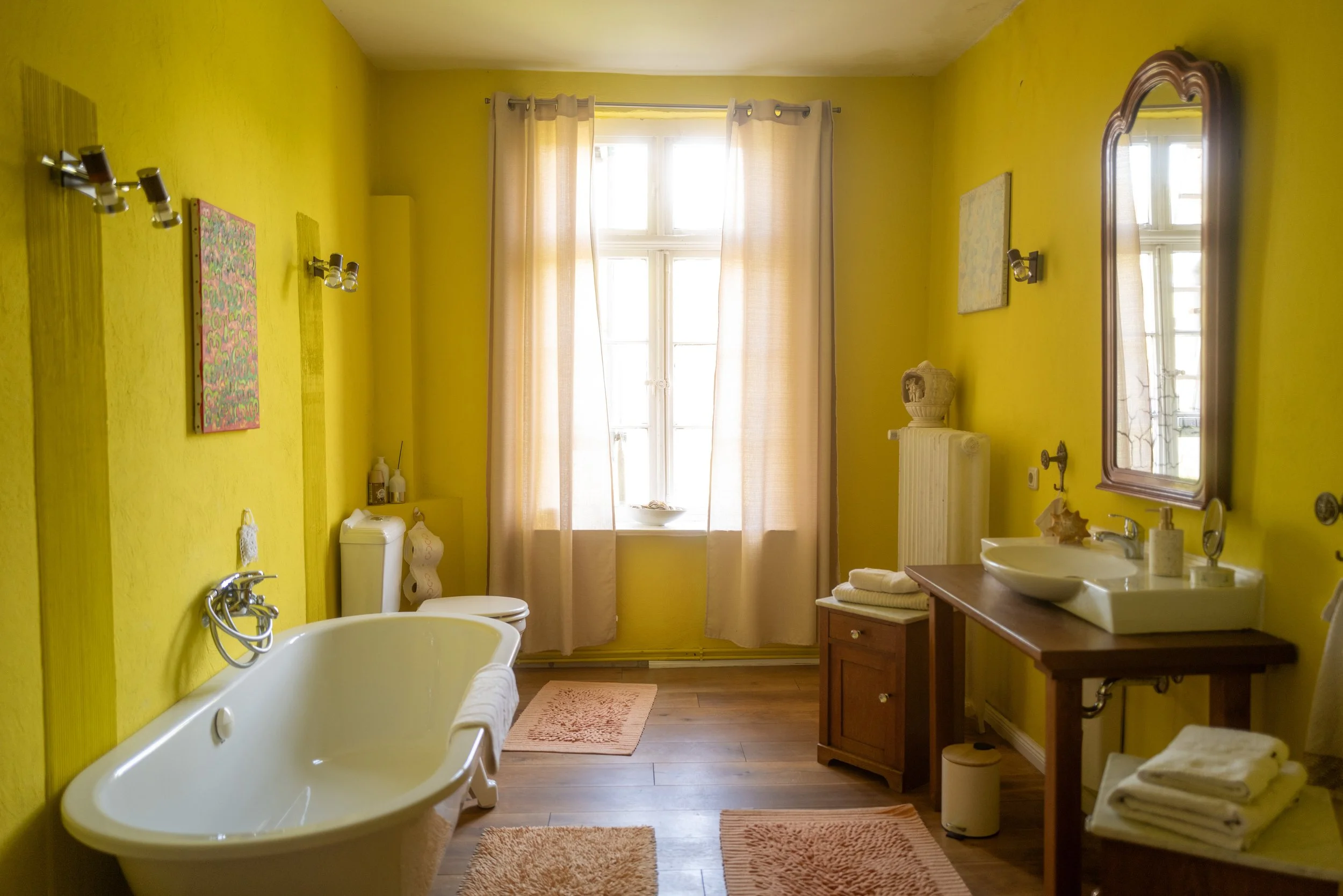 Ein Badezimmer mit gelben Wänden, einer Badewanne, einem Bidet, einem Waschbecken, Spiegel und Vorhängen vor einem Fenster.