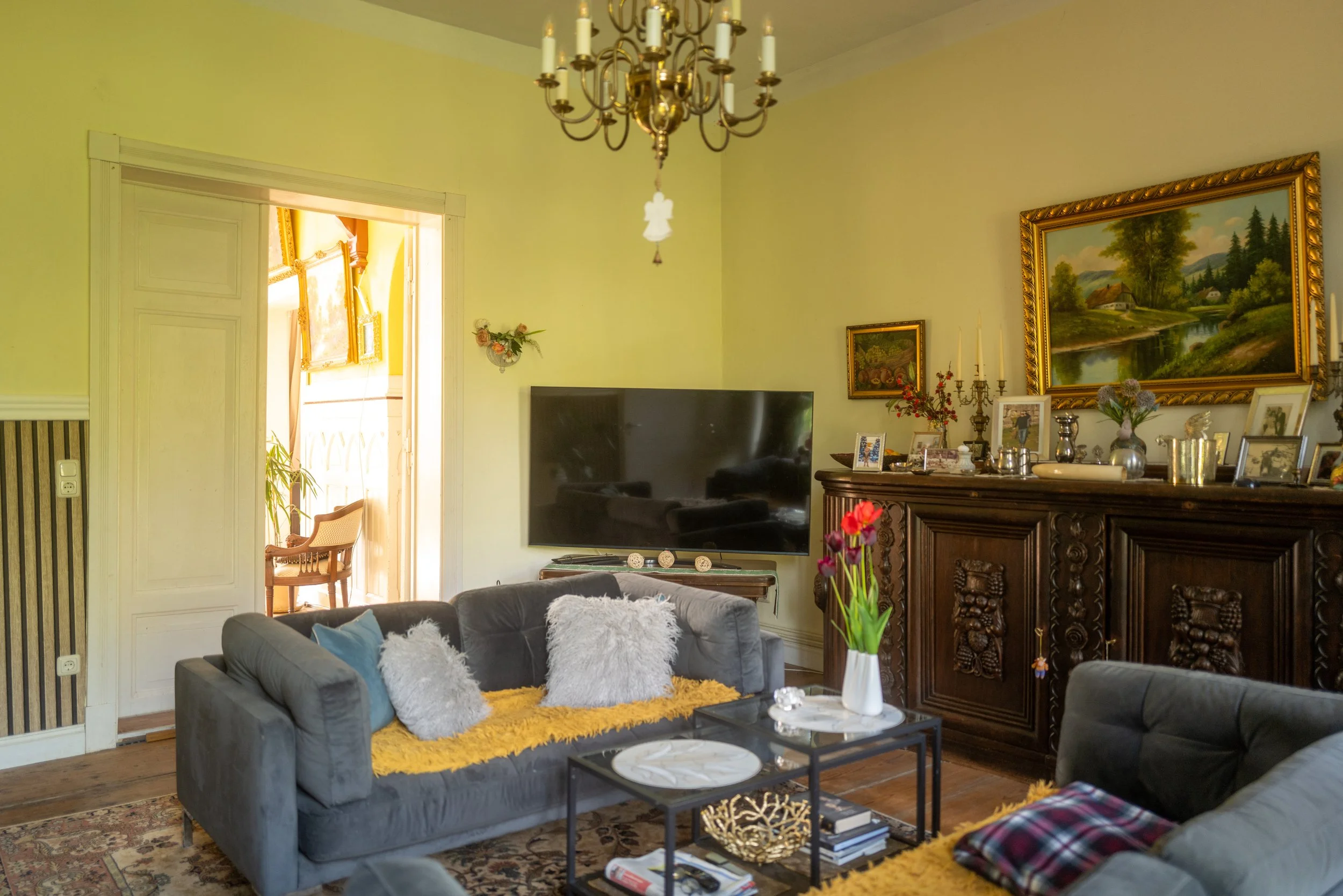 Gemütliches Wohnzimmer mit grauen Sofas, weißen und bunten Kissen, einem braunen Holzsideboard mit Bildern und Dekoration, einem Fernseher, einem Klavier, gehängten Gemälden an der Wand und einem Kronleuchter, viel Tageslicht durch offene Türen.