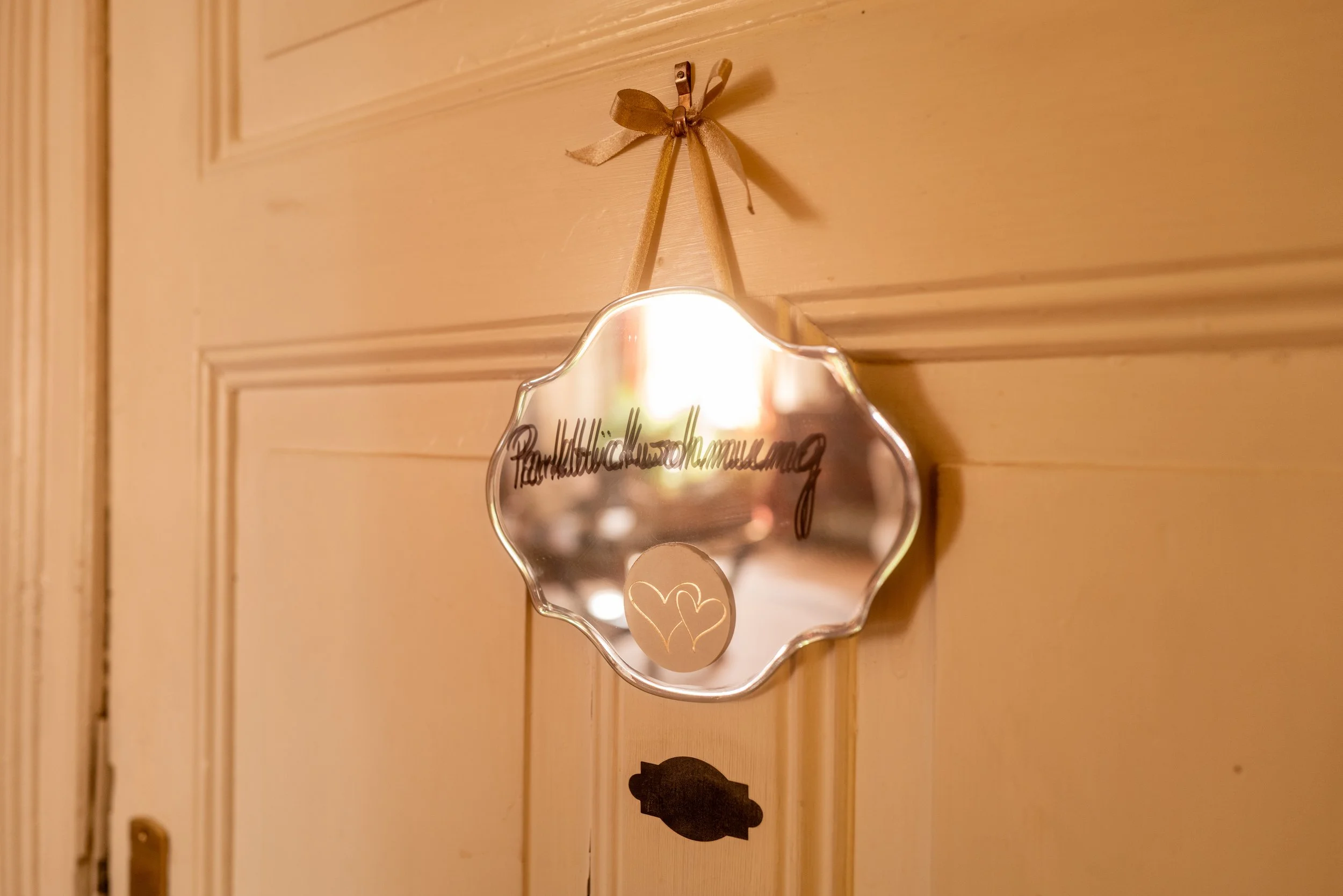 Ein handgeschmiedeter Metallspiegel mit Herzmotiv, der an einer Holzhauttür mit einer goldenen Schleife befestigt ist.