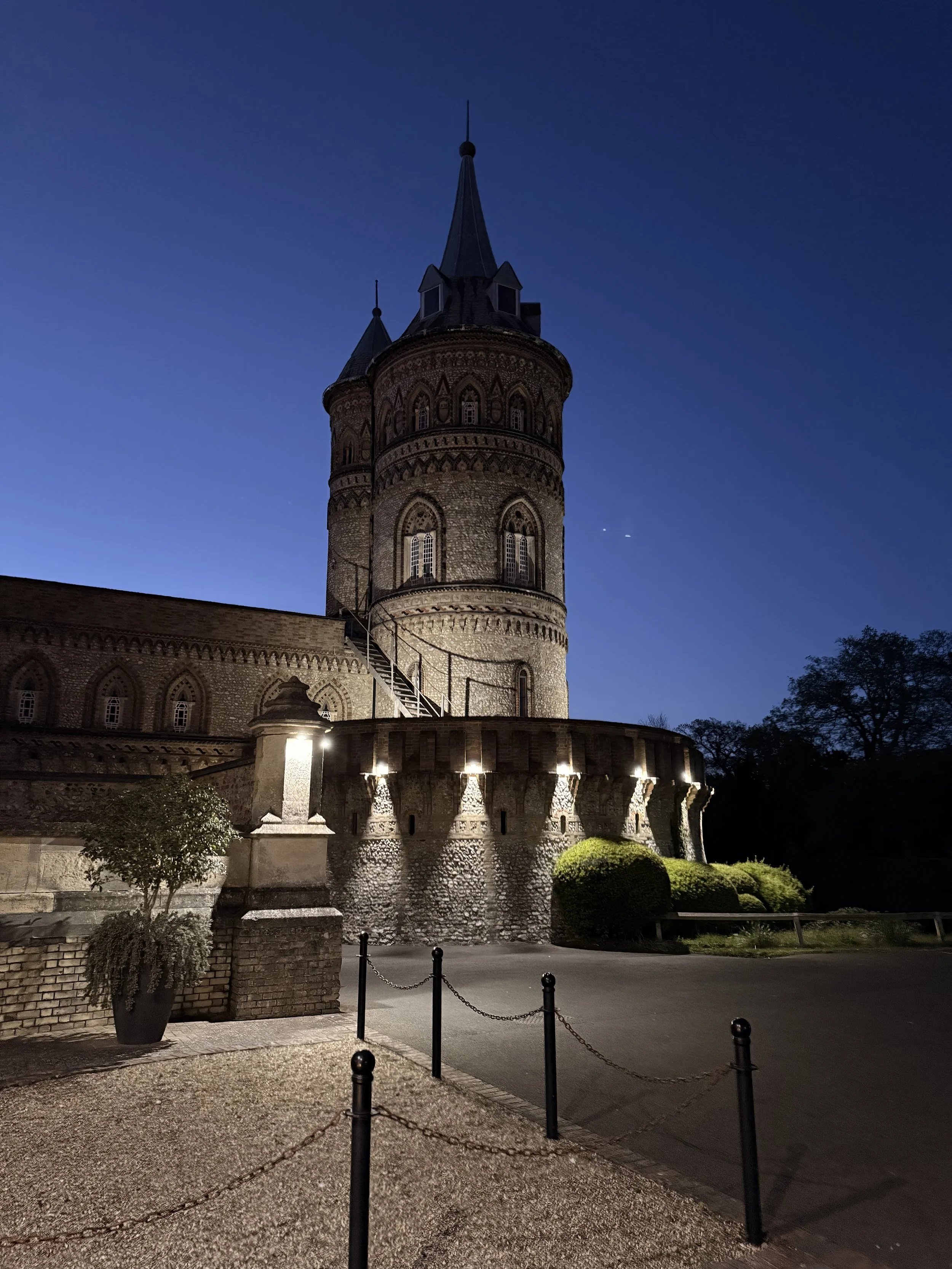 A historic stone castle tower illuminated at night with decorative lights, surrounded by a courtyard with bushes, a potted plant, and a paved pathway with chain barriers.