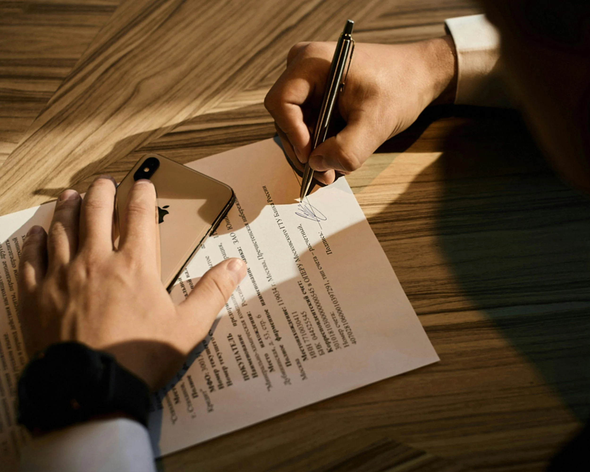 A person signing a document on a wooden table with a gold-colored smartphone nearby.