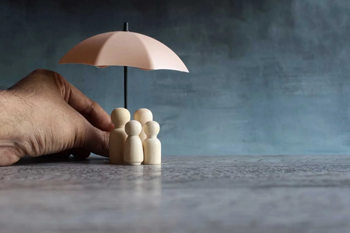 Hand holding wooden figurines of people under a pink umbrella on a wooden surface.