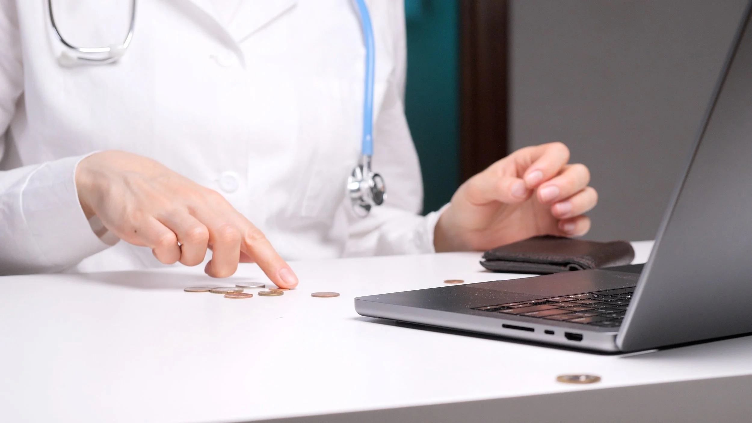 A healthcare professional wearing a white coat and stethoscope counting coins on a white table while working on a laptop.