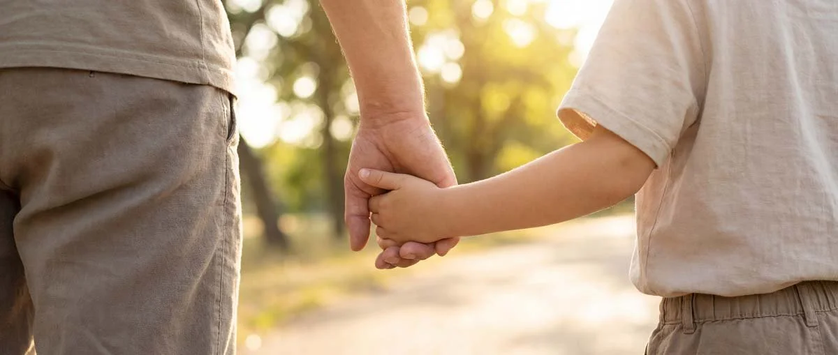 An adult and child holding hands outdoors with sunlight filtering through trees.