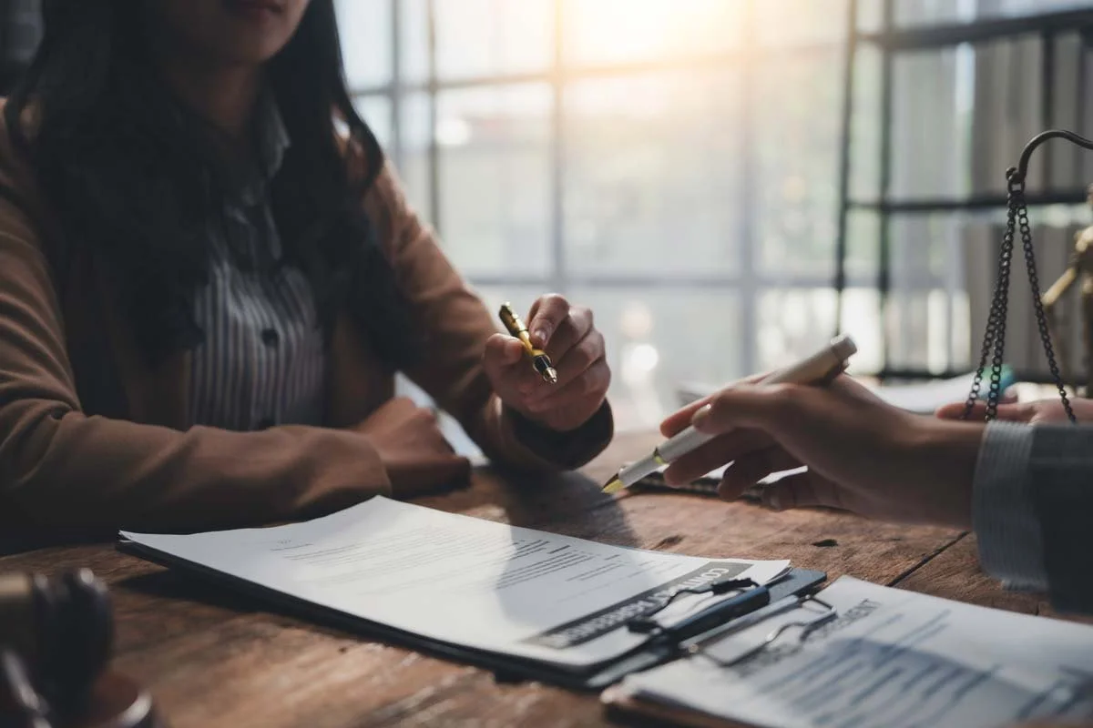 Two people sitting at a wooden table during a meeting, with documents, a clipboard, and pens. One person is holding a pen and gesturing, while the other has a hand extended with a pen.