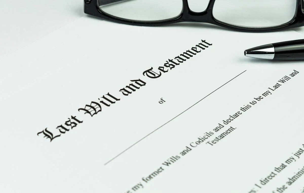 A last will and testament document on white paper, with black eyeglasses, a black pen, and a portion of text visible.