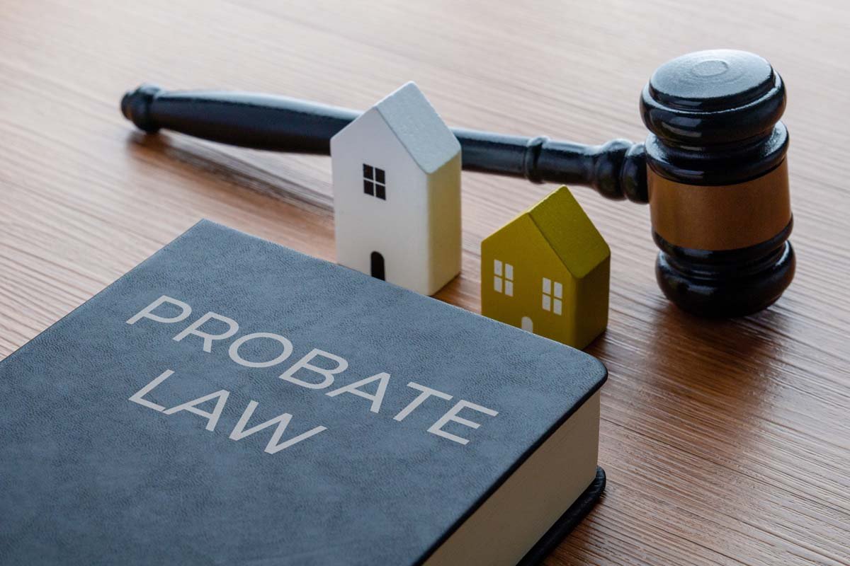 A legal gavel, two small model houses (one white and one yellow), and a book labeled 'PROBATE LAW' on a wooden surface.