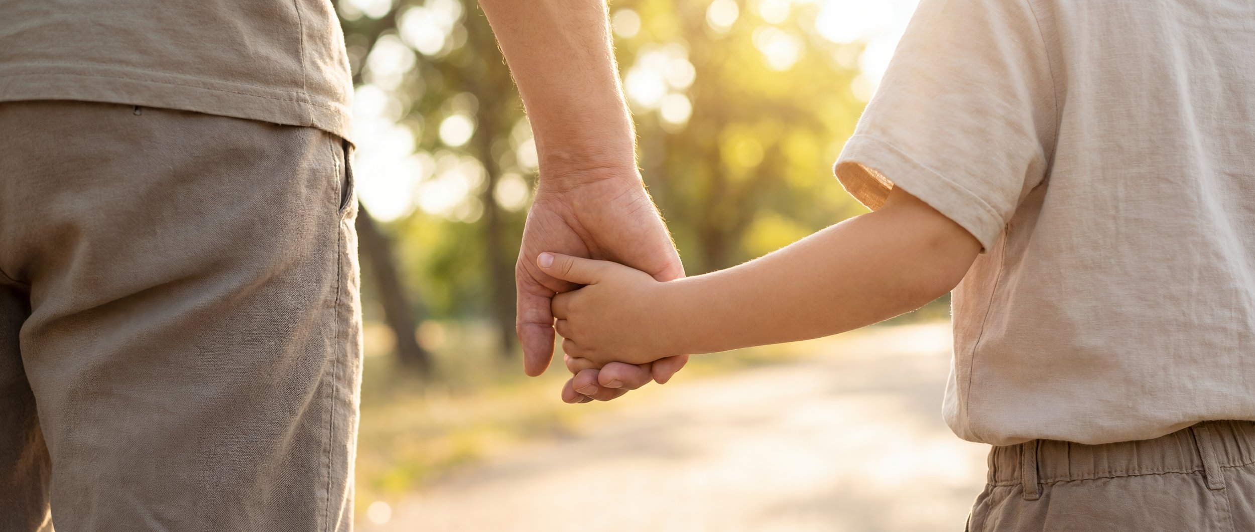 A child and adult holding hands outdoors in a park with sunlight filtering through trees.