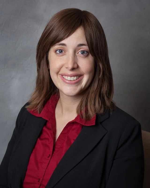Professional woman with shoulder-length brown hair, wearing a black blazer and red shirt, smiling at the camera against a gray background.