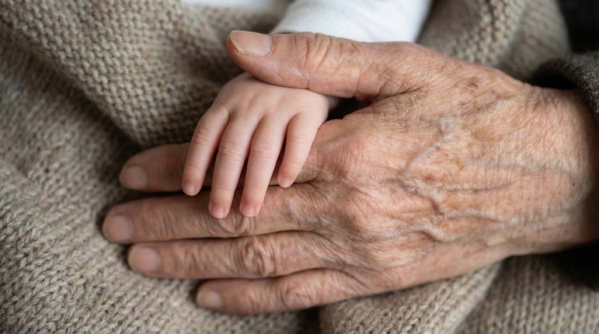 Close-up of an elderly person's hand holding a small child's hand, both resting on a knitted fabric blanket.