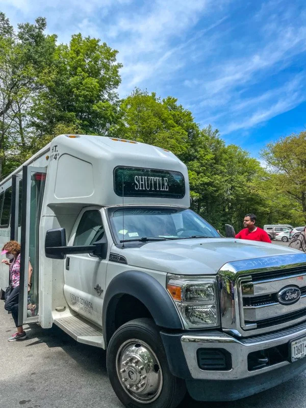 A white shuttle bus with a sign reading 'SHUTTLE' parked in a lot with trees and a blue sky in the background. Two children and an adult are nearby.